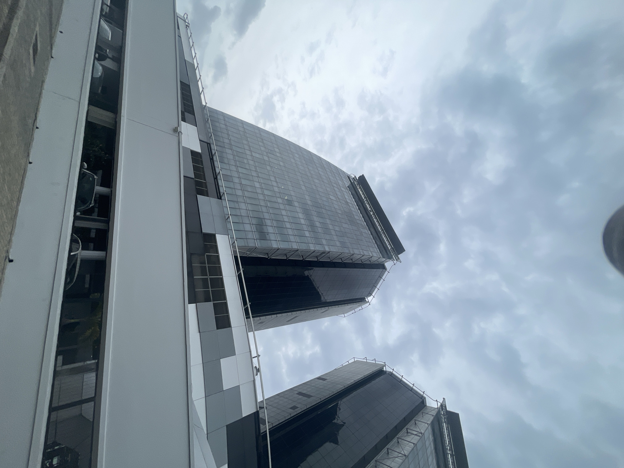 The image shows a low-angle view of tall buildings in Itirin, Nigeria. The buildings are modern, with glass and metal facades. The sky is overcast. There are no people visible. One building has what looks like a parking garage to its left.