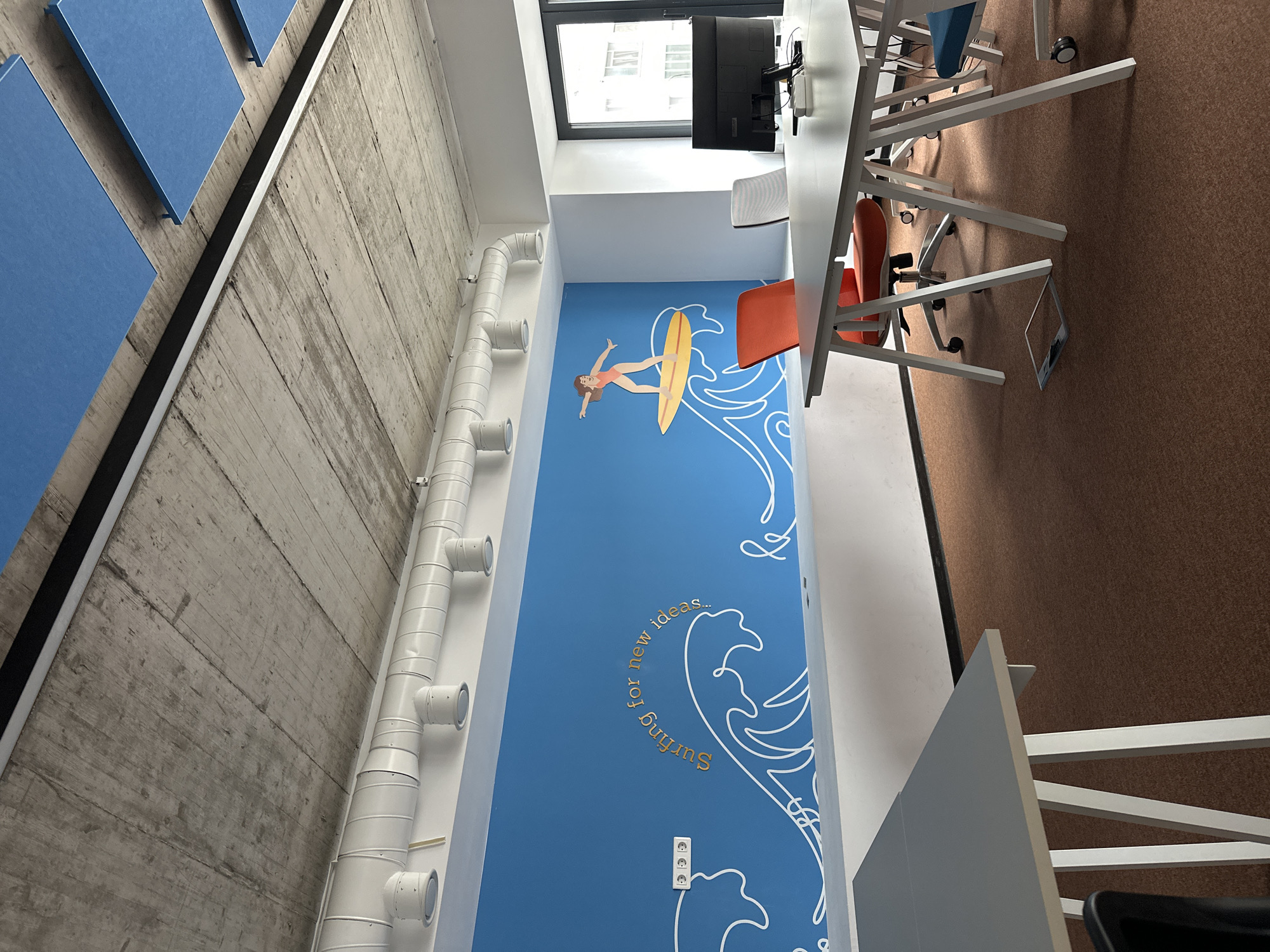 The image depicts an office space with a modern, minimalist design. The color scheme predominantly consists of blue, white, and brown.
On the left side, a concrete wall with horizontal wooden imprints is visible. Attached to this wall are square blue acoustic panels. A series of white pipes runs vertically along the lower part of the wall.
The adjacent wall is painted blue and features a mural depicting a surfer and stylized waves. The text "Surfing for new ideas..." is incorporated into the mural. A power outlet is located near the bottom of the mural.
A long white table runs along the right side of the image. Several office chairs, primarily in white and orange, are positioned around the table. A computer monitor is mounted on the wall above the table.
The floor is covered with brown carpet.
A window is visible at the top of the image, suggesting natural light entering the space.
The specific location in Varna, Bulgaria, cannot be determined from the given image.