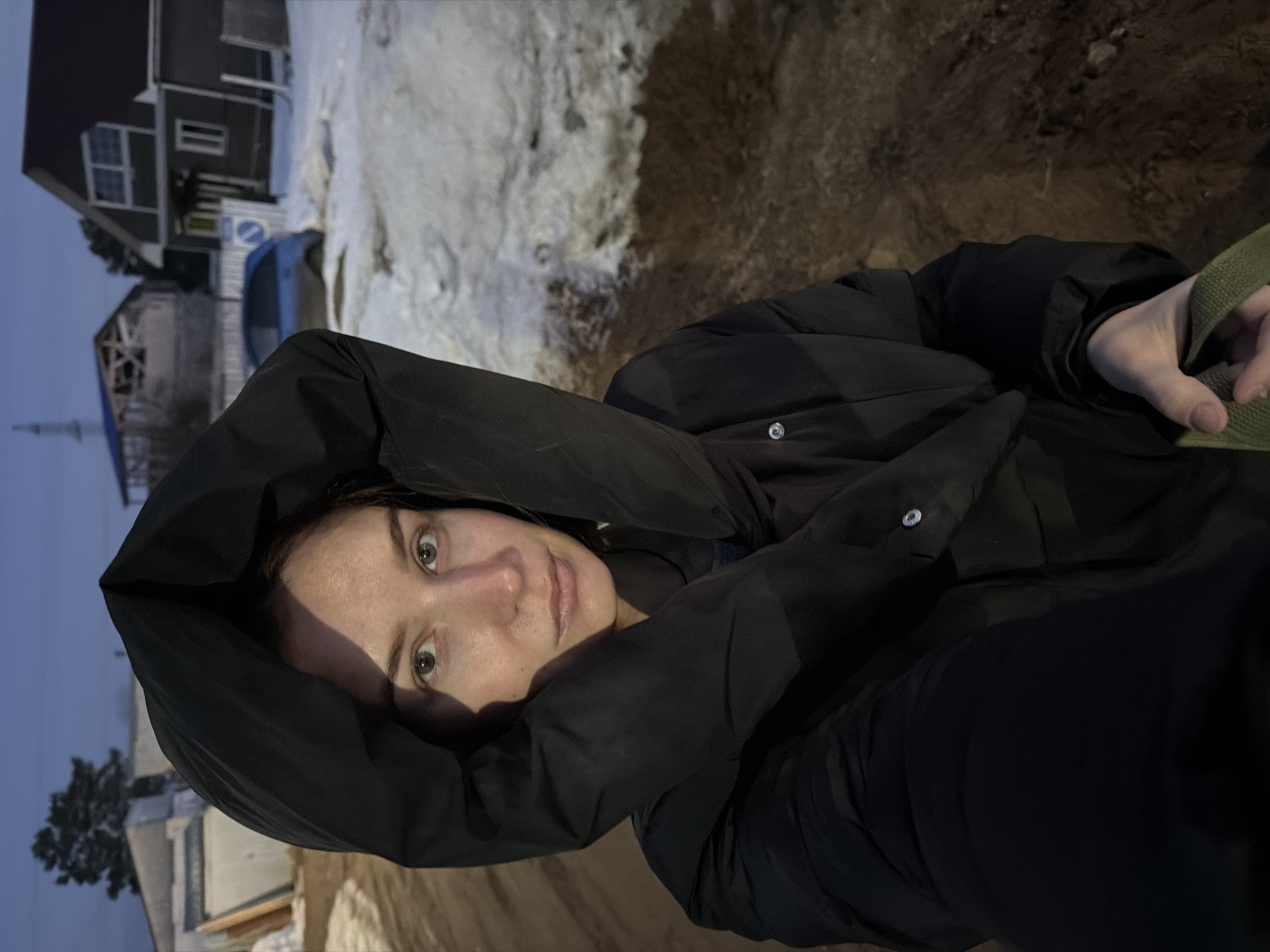 The image is a selfie of a woman wearing a black hooded jacket, looking directly at the camera. The background reveals a snowy landscape, likely in a residential area. A house is visible in the upper left corner of the frame. Based on the location information provided, this image was likely taken in Chaikovsky, Russia. The scene appears to be outdoors, possibly during dusk or dawn, given the lighting. The woman has fair skin, light eyes, and her hair is tucked under the hood. The focus is sharp on her face. She is holding a green object (possibly a bag strap) in her right hand.