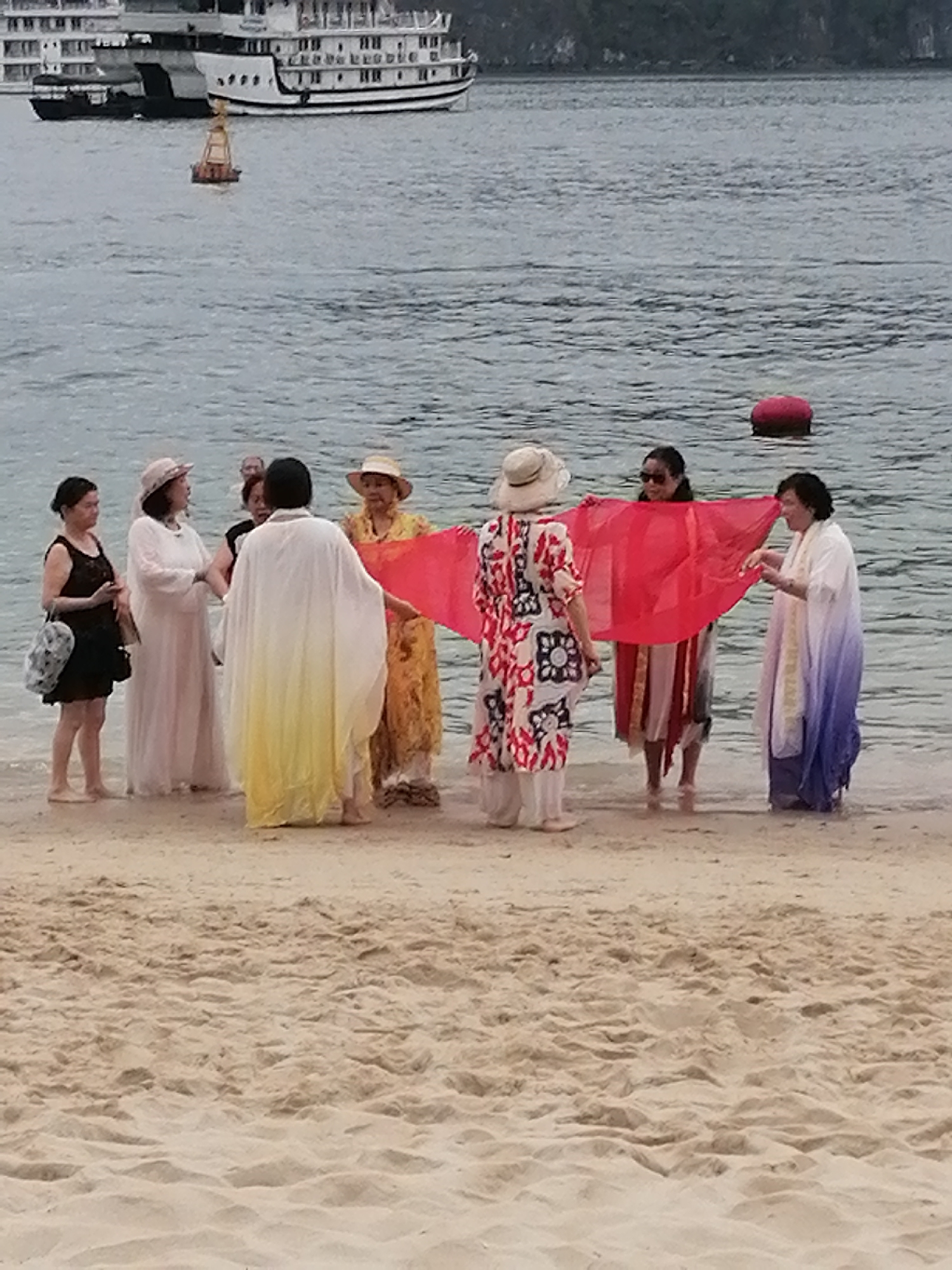 The image depicts a group of approximately eight to nine women gathered at the sandy edge of a beach in Ha Long Ward, Vietnam. The scene is set on a seemingly overcast day, with calm, grey-green water meeting a light brown sandy shore.
The subjects are predominantly women, most of whom are dressed in loose-fitting, resort-style or traditional attire. Their clothing includes long, flowy garments in various colors such as white, a white-to-yellow gradient, a yellow patterned dress, a red and white patterned tunic, and a white-to-purple gradient outfit. Several women wear wide-brimmed hats, and most are barefoot or in sandals, standing in the shallow water or on the wet sand.
The primary activity involves a central group of women holding up a large, vibrant red sheer fabric, which they are spreading open. This action suggests they are posing for a photograph or creating a visual display with the fabric. Other women stand nearby, observing or interacting with the main group.
Notable details include a large, white multi-deck cruise ship or tour boat anchored in the distance to the left, and a smaller yellow-orange buoy floating closer to the vessel. Another red buoy is visible further out in the water on the right. The background features distant, dark landmasses or cliffs covered in greenery, typical of the Ha Long Bay landscape. The sandy foreground shows ripples and textures created by the receding tide. The overall atmosphere appears leisurely, suggesting a vacation or group outing. There is no visible text in the image.