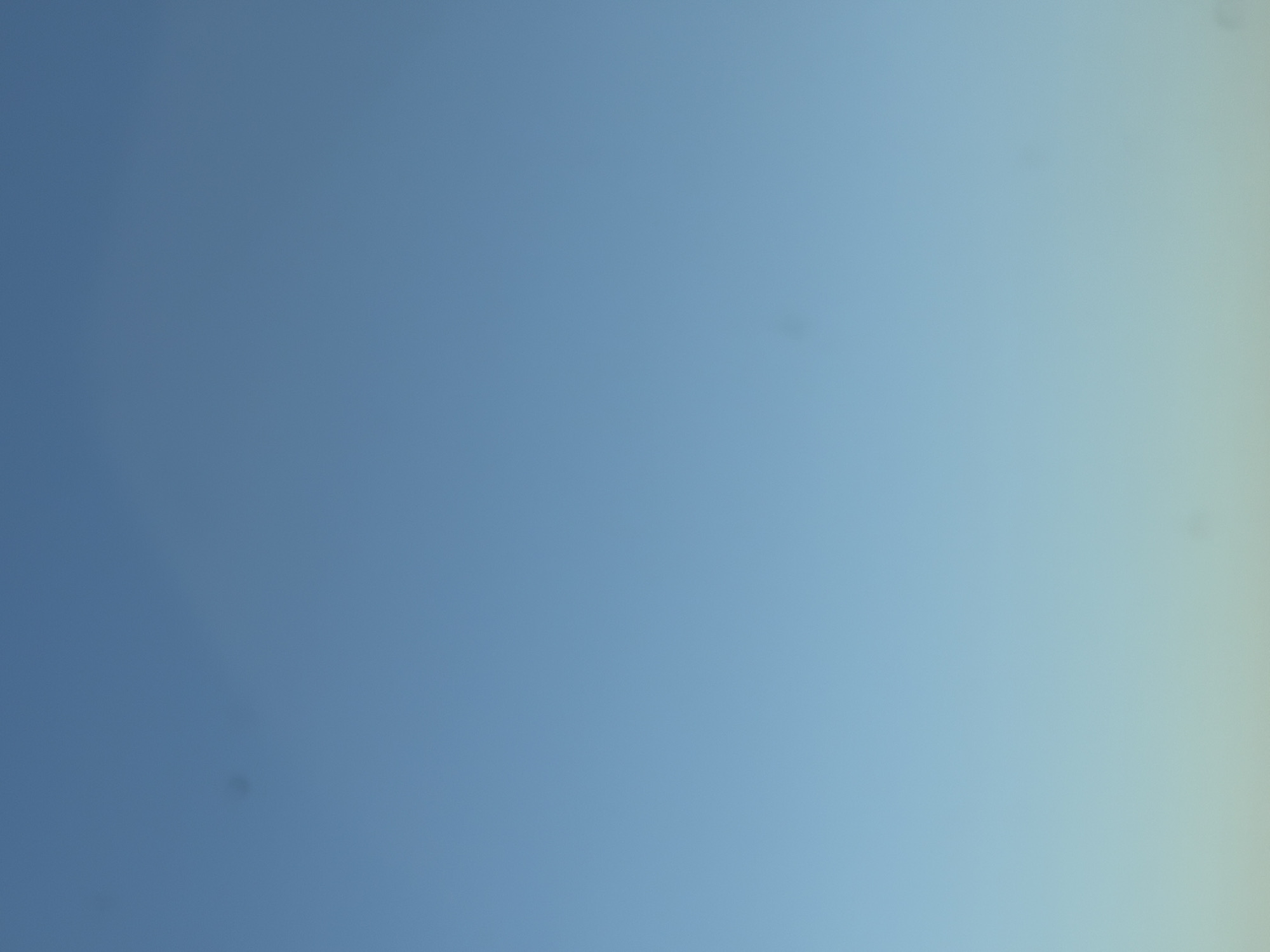 The image is a photograph of a clear blue sky with a slight gradient towards a lighter shade at the bottom. There are a few small, dark circular blemishes present on the sky, likely dust spots on the camera lens. No people, subjects, objects, actions, or interactions are visible. The image does not provide any identifiable landmarks or context to determine its location in Sereď, Slovakia.