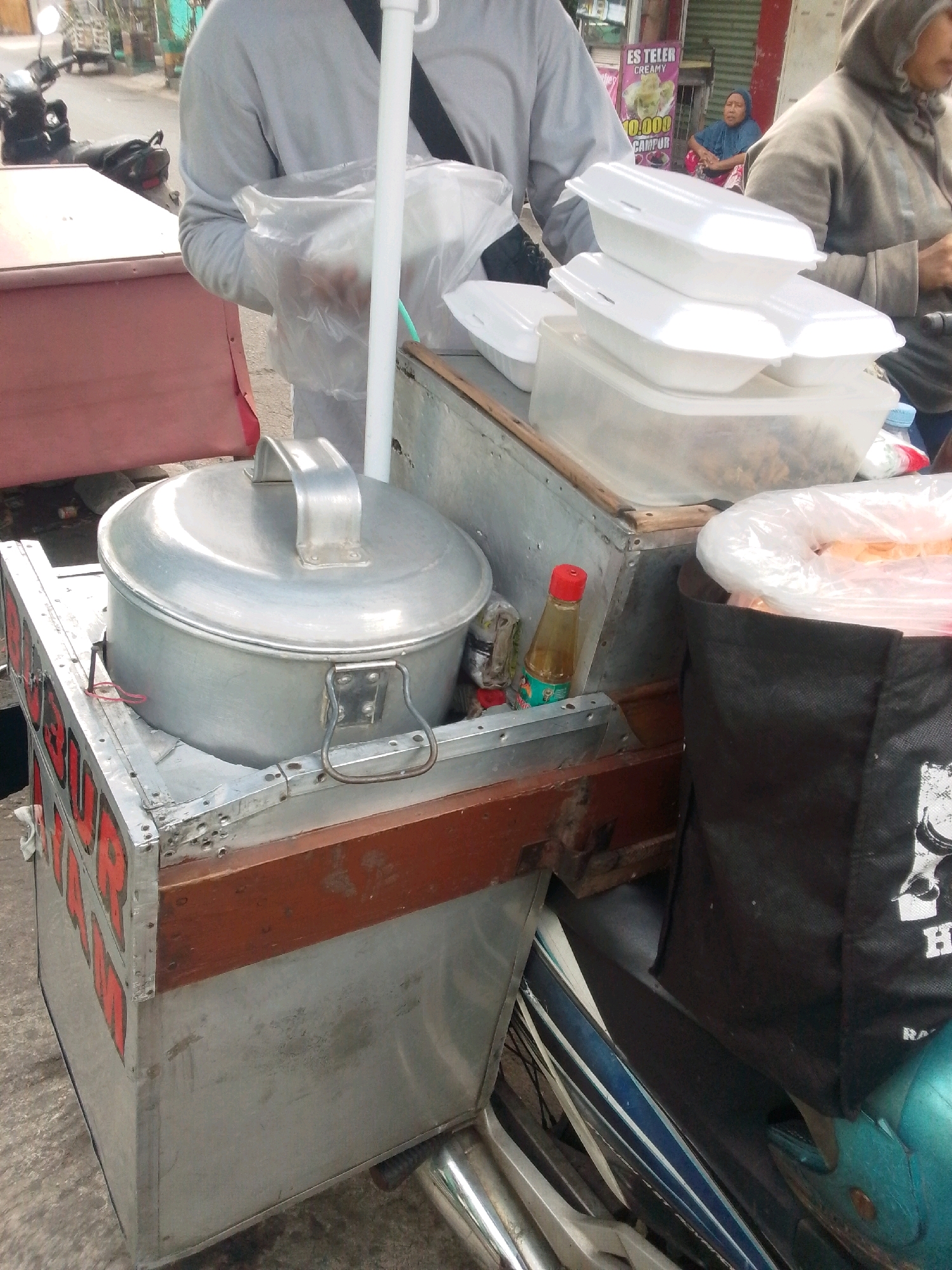A street food cart is pictured on a sidewalk in East Jakarta, Indonesia. The cart is made of metal and wood, and it contains various food items and containers. A large silver pot with a lid is prominently displayed on the cart, along with a stack of white styrofoam food containers. There is also a black bag, a bottle of sauce, and a plastic bag filled with food. A person, wearing a gray long-sleeved shirt and holding a plastic bag, is standing behind the cart. Another person, wearing a gray hooded jacket and a blue headscarf, is seated behind the cart. In the background, there is a sign that reads "ES TELER CREAMY" and a motorcycle parked nearby. The ground is made of concrete.