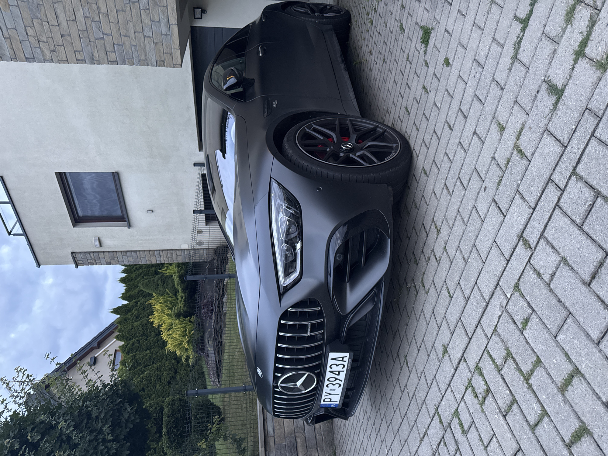 A matte black Mercedes-AMG GT sedan is parked on a herringbone brick driveway in front of a modern house. The house has white stucco walls and a stone-clad section. The car's front grille features vertical chrome slats and a prominent Mercedes-Benz star emblem. The license plate reads "PY 3943A". Red brake calipers are visible behind the black multi-spoke wheels. Lush green trees and shrubs form a backdrop, with a wire fence separating the property from the street. The sky is overcast.