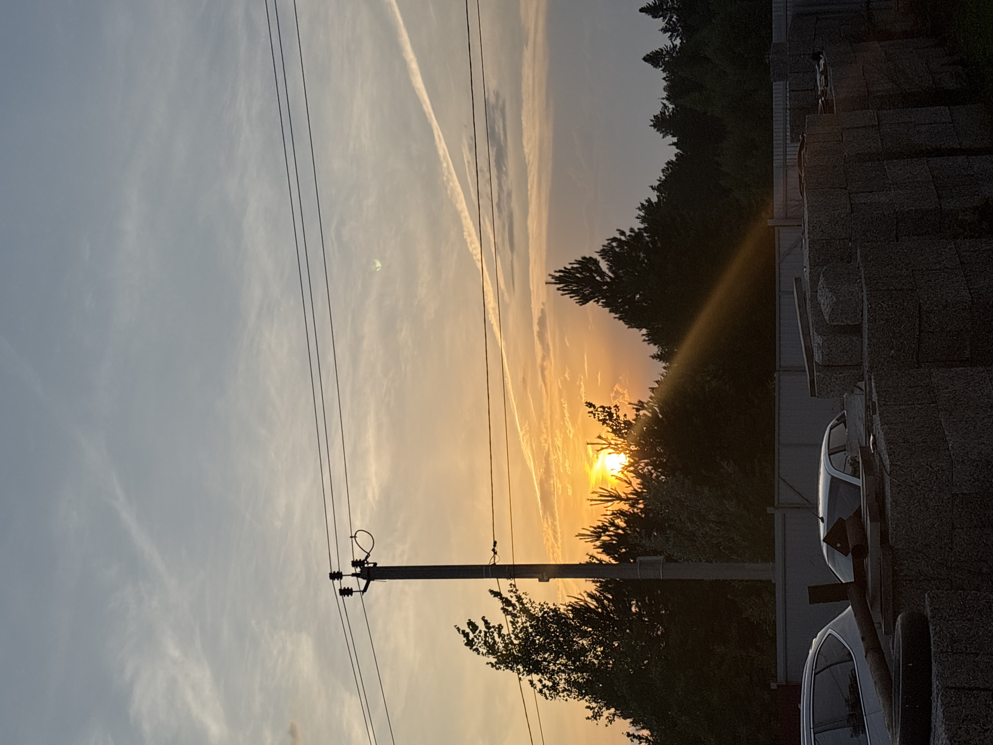 The image depicts a sunset over a rural landscape in Прикамский, Russia. The sun, a bright orb, is partially obscured by trees and streaks of clouds, casting a warm, orange glow across the sky. Several power lines stretch across the frame, supported by a utility pole on the left side of the image. In the background, a dense line of trees forms a dark silhouette against the vibrant sky. On the right side of the image, a stone wall or structure is visible, with two cars parked alongside it. One car is white, and the other appears to be dark. The overall scene evokes a sense of tranquility and the peaceful end of a day. The presence of power lines and cars suggests a populated area, likely a village or small town. The weather appears clear, with the exception of some wispy clouds that add texture to the sky. The time of day is clearly sunset, indicated by the low angle of the sun and the warm color palette. There is no visible text in the image.