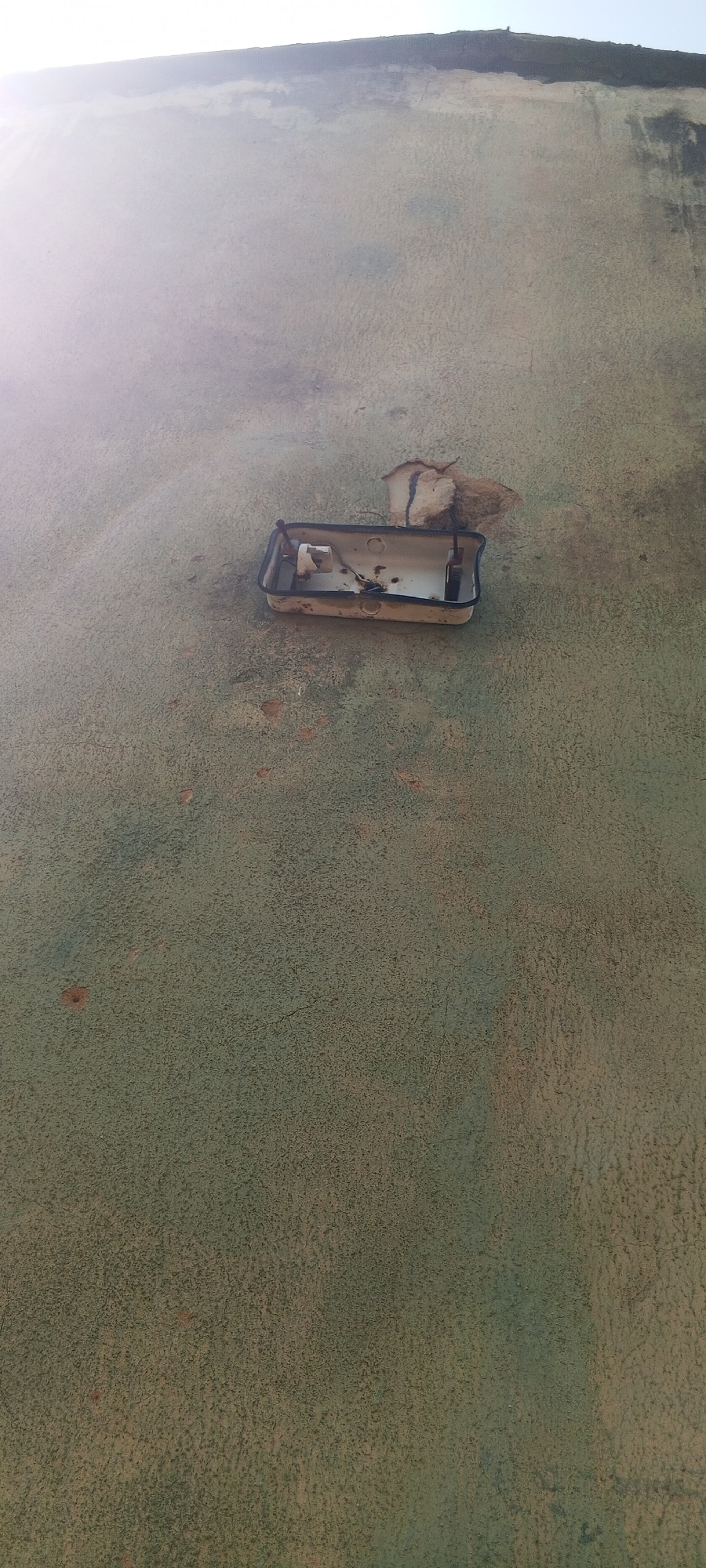 The image depicts a close-up, vertical shot of a textured, greenish-grey wall in Jalingo, Nigeria. A detached, rectangular, white fixture with a black rim is affixed to the wall. The fixture appears to be a light housing, possibly for an outdoor light, and it is empty and damaged. The surface of the wall exhibits a rough, speckled texture with visible signs of wear and some small, circular depressions. In the upper portion of the image, a bright, washed-out sky is visible, with a darker, horizontal line suggesting the edge of a roof or structure. The lighting suggests it is daytime, with the sun possibly behind the camera or to the left, creating a bright, hazy effect in the upper left quadrant of the image. There are no people or animals visible in the frame.