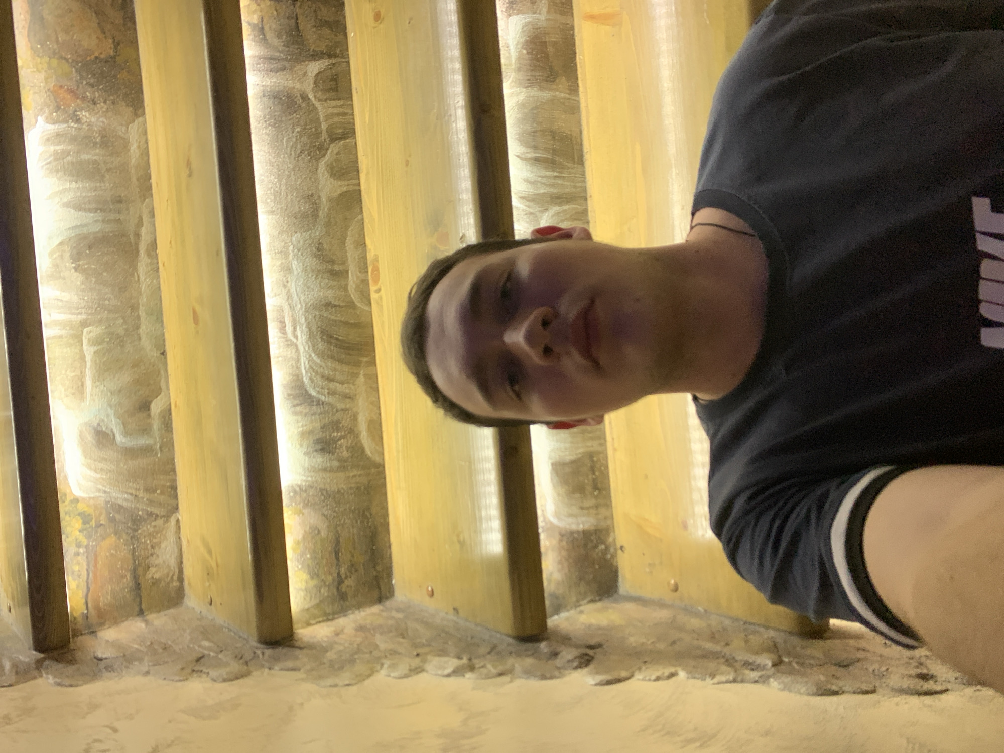 A man wearing a dark Nike t-shirt stands in front of a decorative wall, which consists of vertical wooden planks and a textured, possibly stone-like, filling between the planks. The wall is lit with a warm, possibly artificial light. The man is looking directly at the camera. The location is Severodvinsk, Russia.
