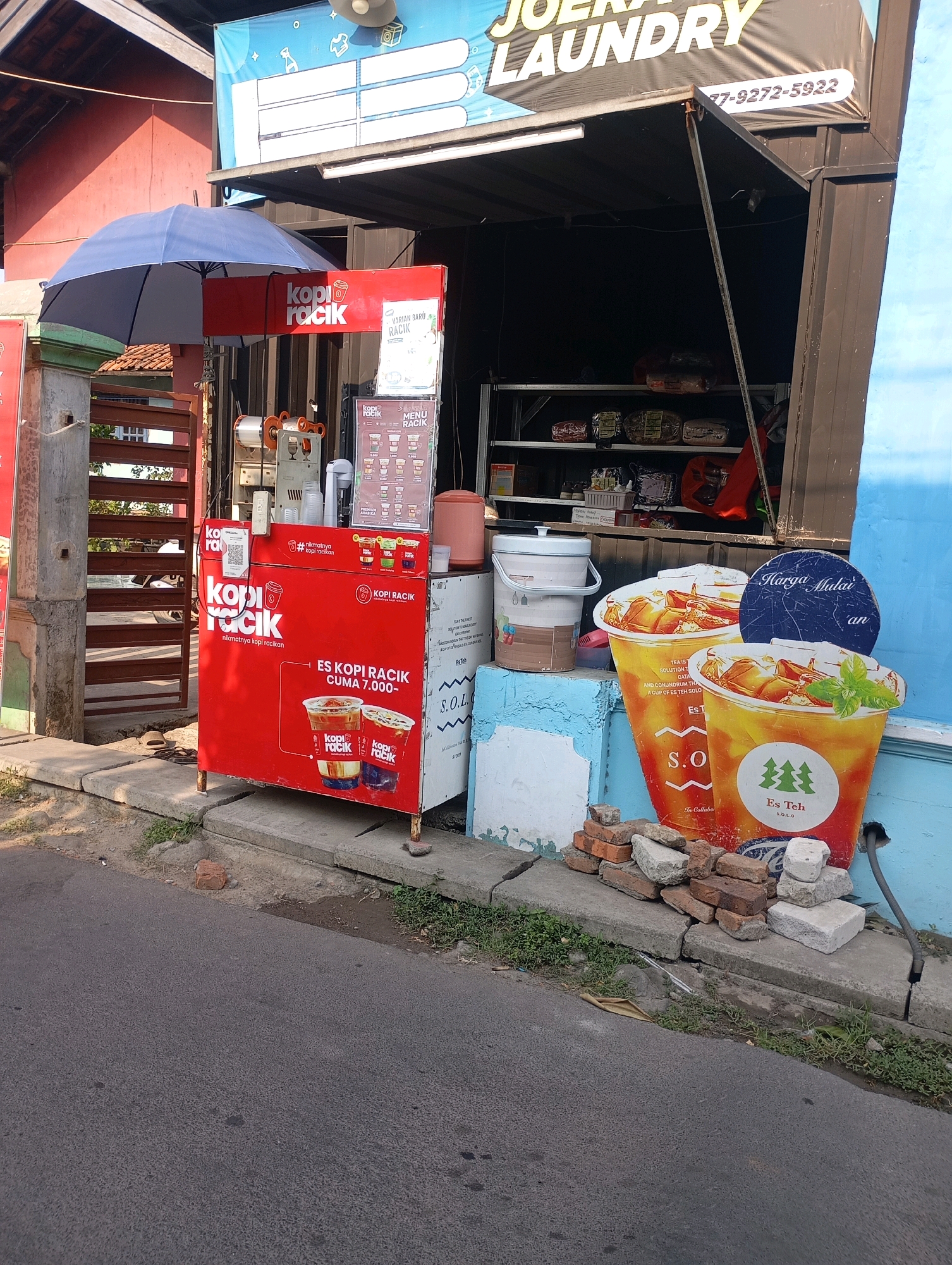 The image shows an outdoor food stall, likely a kiosk, selling coffee and tea. The stall is bright red with "Kopi Racik" (Mixed Coffee) branding prominently displayed. There are menus and images of iced coffee and iced tea on the stall. A large blue umbrella is set up over the stall. In the background, a building with signage for "JOERA LAUNDRY" is visible. The stall is situated on a sidewalk next to a paved road. Scattered around the base of the stall are bricks and building materials. The environment appears to be a sunny day. The specific location is identified as Babelan, Indonesia.