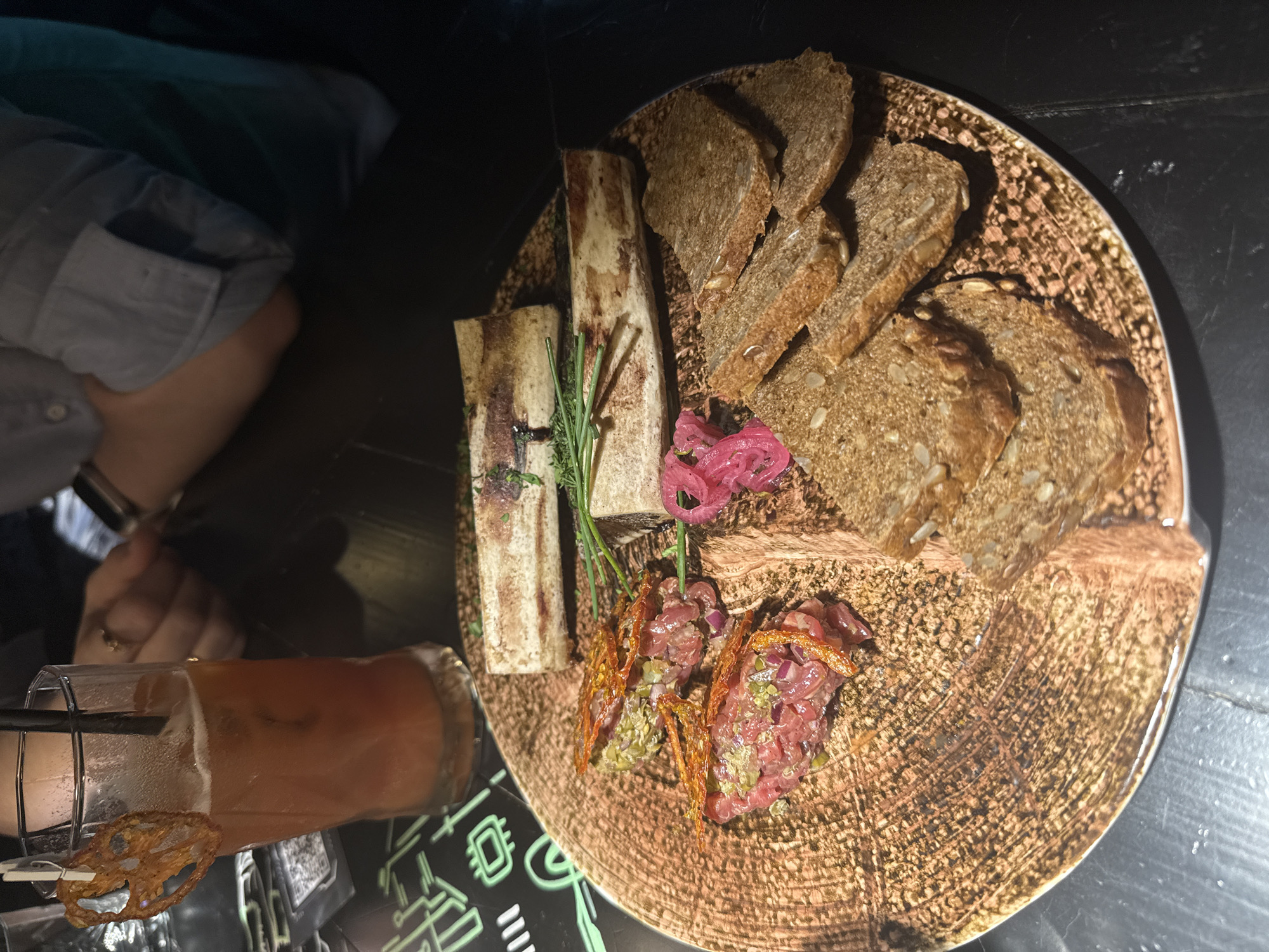 The image shows a food presentation on a textured brown plate. The plate contains marrow bones, slices of seeded bread, pickled onions, and two portions of what appears to be steak tartare, garnished with fried elements. Next to the plate is a glass of beverage with a straw and a dried lotus root garnish. A person is visible on the left side of the frame, wearing a grey shirt, with a ring on their finger. The location is in Saint Petersburg, Russia, based on the presence of a menu or coaster displaying symbols associated with the city. The setting appears to be a dimly lit restaurant or bar, with the food presentation as the main subject.