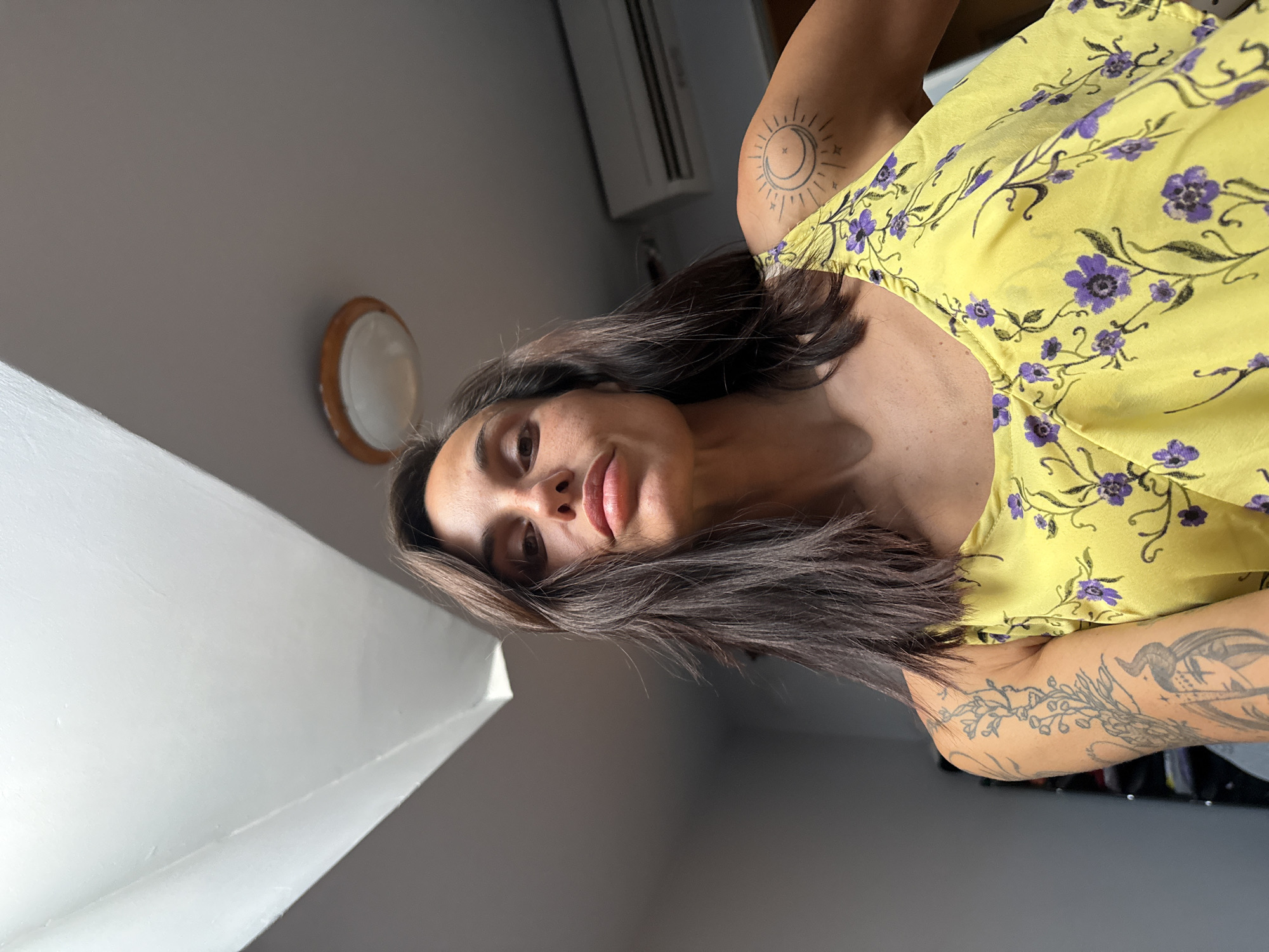 A woman is depicted in an indoor setting, in the city of Pleven, Bulgaria. She has dark brown, medium-length hair and is looking downwards directly at the camera with a neutral expression. She is wearing a yellow sleeveless top with purple floral and green vine patterns. On her right shoulder, a tattoo of a stylized sun and crescent moon is visible. Her left arm displays multiple tattoos, including an intricate botanical vine design and a figurative image featuring a face with horn-like elements. The background consists of a light gray wall and a white ceiling with angular contours. A round ceiling light with a light-colored rim is mounted on the ceiling towards the upper right of the frame, and a white wall-mounted air conditioning unit is visible on the wall behind her right shoulder. The image is taken from a low-angle perspective, looking up at the subject.