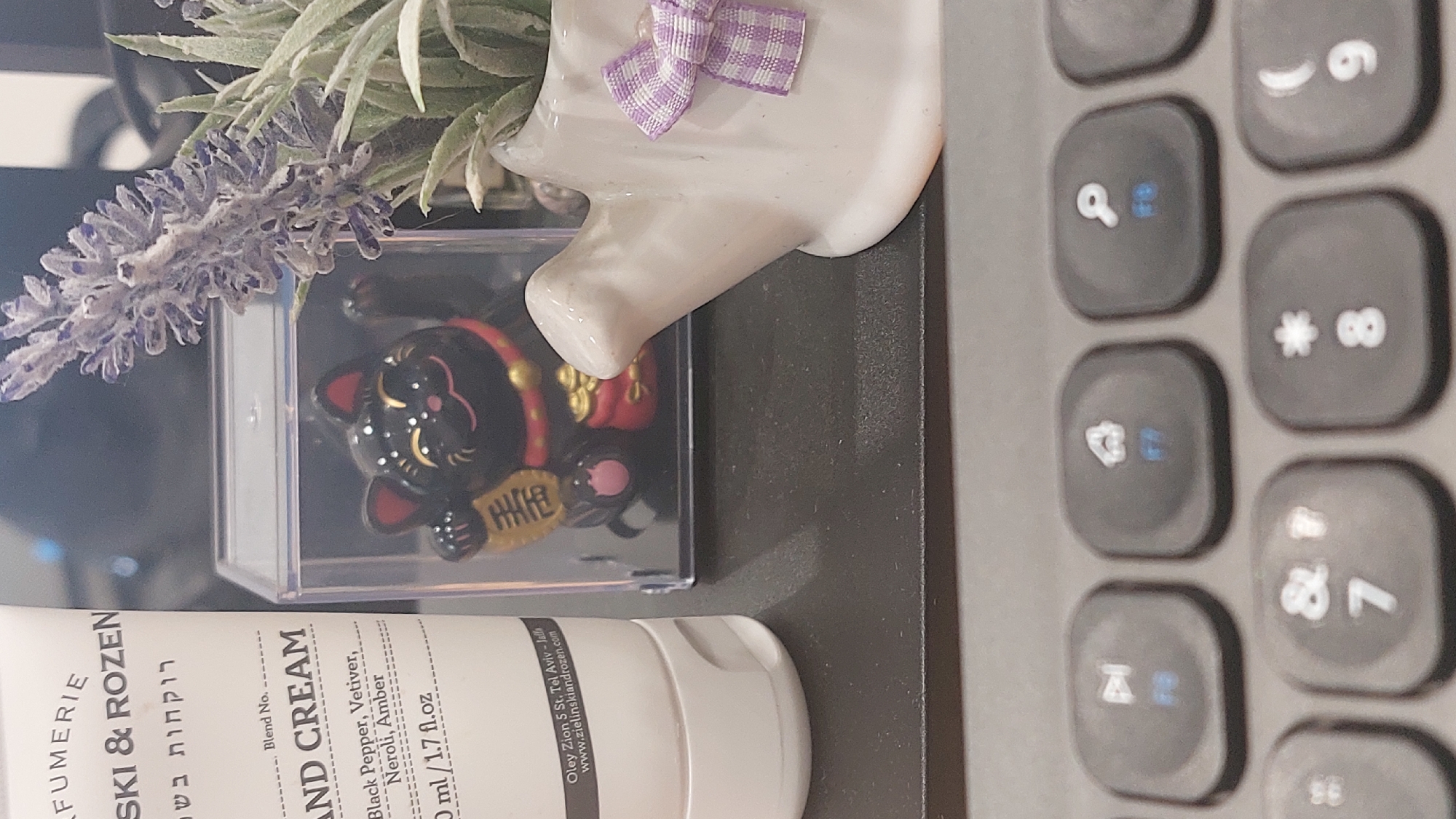 Here is a detailed description of the image:
**Scene:** A close-up, rotated view of a desktop setting.
**Objects:**
* **Keyboard:** A section of a gray keyboard with visible number keys.
* **Hand Cream:** A white tube of hand cream. The brand is visible with the name "Zielinski & Rozen", along with descriptors like "Blend No.", "Hand Cream", and ingredients like "Black Pepper, Vetiver, Neroli, Amber". The tube indicates a volume of "50 ml/1.7 fl. oz". An address for Oley Zion 5 St, Tel Aviv-Jalla is also visible.
* **Ornament/Figure:** A small black cat figure, possibly a Maneki-neko, is visible within a clear case.
* **Plant:** A sprig of artificial lavender, and other artificial green leaves.
* **Vase:** A small, white vase with a purple and white checkered bow.
**Environment:**
* The general color scheme is muted, consisting of grays, whites, and some muted greens and purples.
* The setting is a desk surface with a computer or laptop nearby, given the presence of the keyboard.
There are no people present in the image. The location in the city of Moscow, Russia, is not shown.