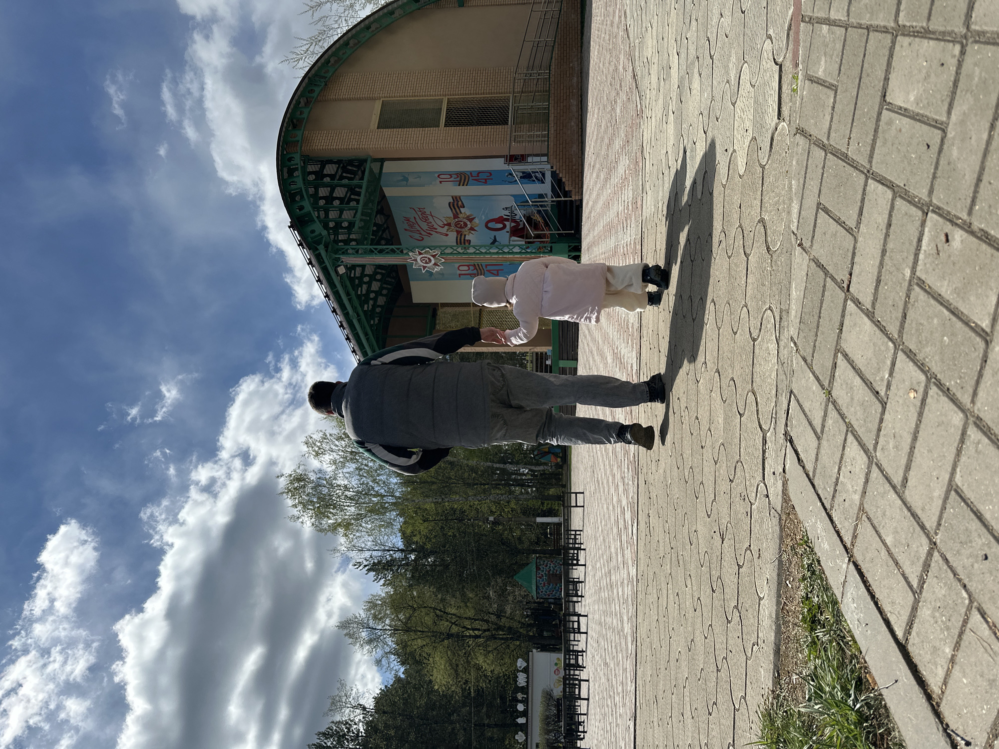 A waist-up view of an adult male and a toddler, both seen from behind. They are standing on a paved surface in the city of Chaikovsky, Russia, in front of a building decorated with Soviet-era imagery commemorating Victory Day (May 9th). The toddler, dressed in a light-colored outfit and a hat, appears to be handing something to the adult. The adult, wearing a dark jacket and light-colored pants, reaches out to receive it. Behind them is a park with trees. The sky is partially covered with clouds.