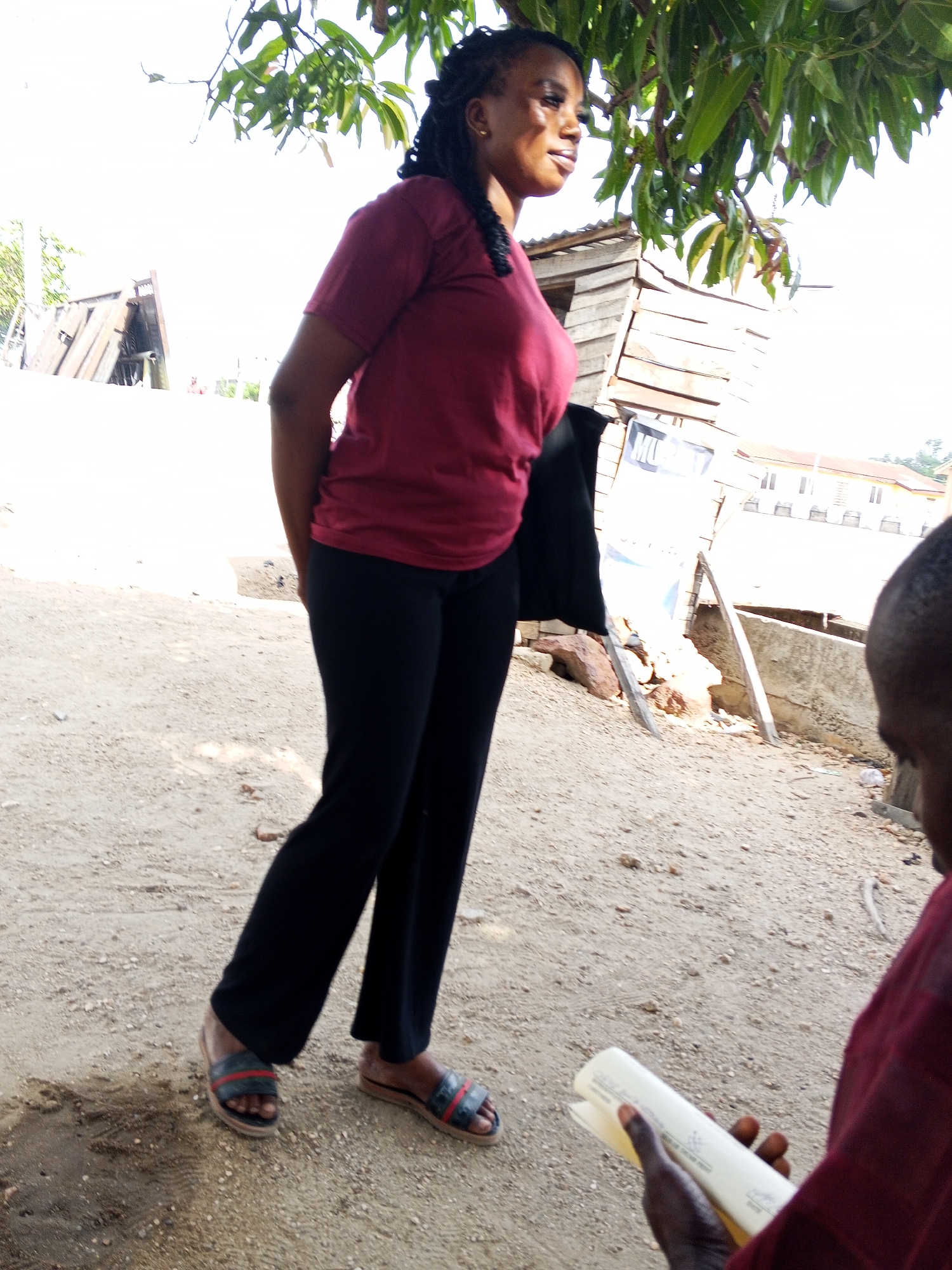 The image shows a woman standing on a dirt road in Zango-ndaji, Nigeria. She is wearing a maroon t-shirt, black pants, and sandals. She has dark braids. A black jacket is hanging over her left shoulder. To the left of the woman is a wooden structure. A poster is held by wooden stakes to the right of the woman. A person in a maroon shirt is holding a white object in the lower right corner. A tree is visible at the top of the frame. The environment appears to be outdoors.