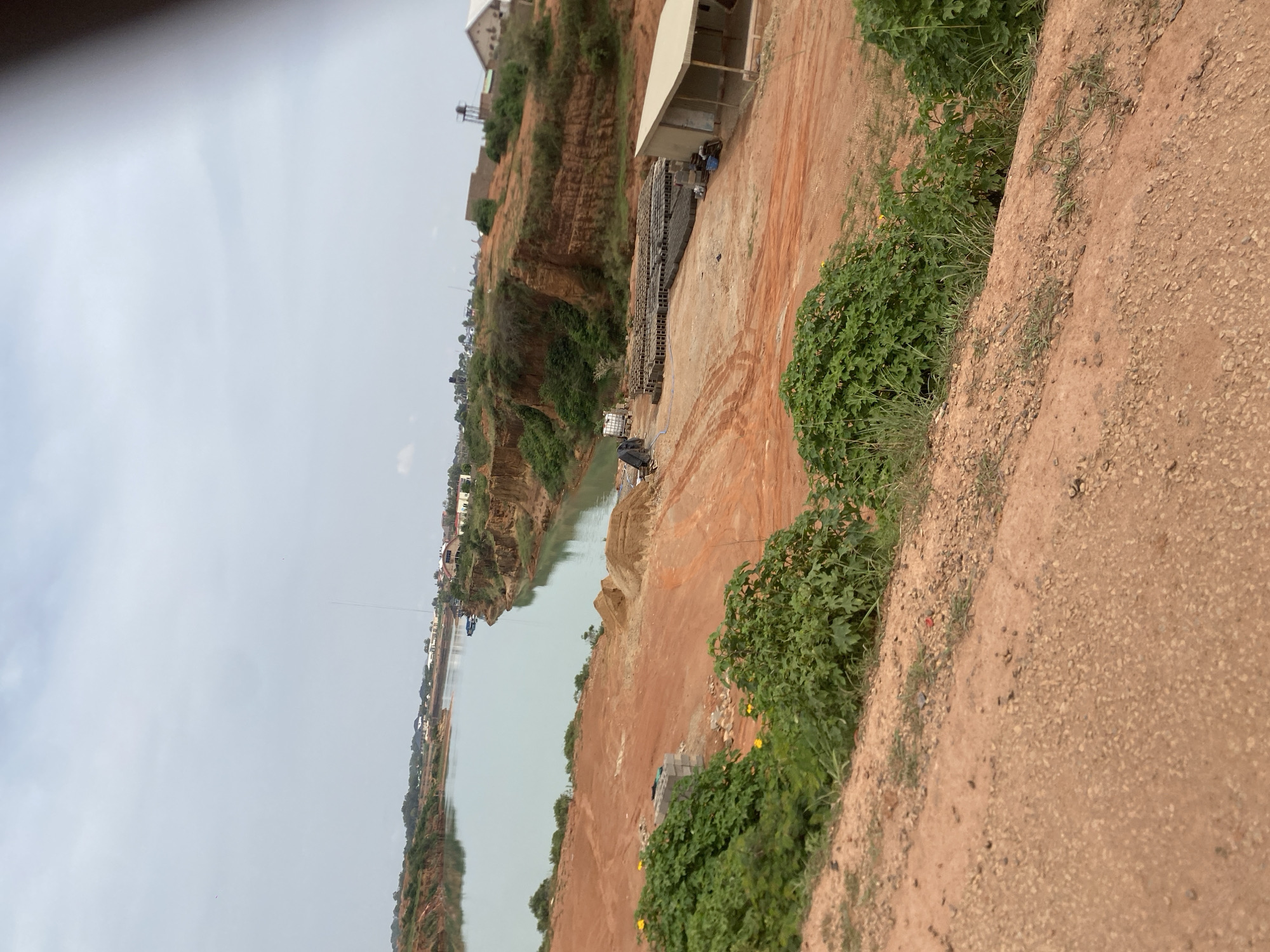 An aerial, slightly blurred view looks down upon a body of tranquil, light blue water, possibly a quarry lake, bordered by steep, reddish-brown earthen cliffs. The cliffs are sparsely covered with green vegetation, and the top of the cliffs are dotted with buildings. In the foreground, the landscape is a barren, reddish-orange dirt expanse, with mounds of dirt and piles of grey blocks. Several green bushes with broad leaves dot the foreground. In the middle ground, a cluster of grey blocks is arranged in rows, and a small, light-colored building with a slanted roof stands nearby. A white truck is parked to the left of the blocks. In the distance, a tall, slender tower pierces the overcast, grey sky. The overall scene suggests a construction or excavation site near a body of water, with residential buildings visible in the background.
