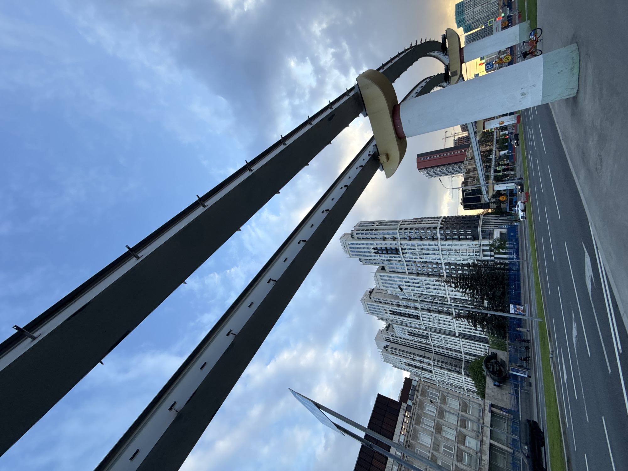The image depicts a street scene in Khimki, Russia, under a cloudy sky. Prominently featured are two curved gray steel structures supported by large, cylindrical white pillars with cream-colored sections, likely an architectural element or artwork above the road. In the background, there are several tall, modern apartment buildings with white facades. A road with lane markings runs alongside the buildings, showing a mix of pedestrian walkways, vegetation, and traffic. There is also a possible monument or statue along the sidewalk. The perspective is angled upwards, emphasizing the height of the structures and buildings. A bicycle is visible on the edge of the image.