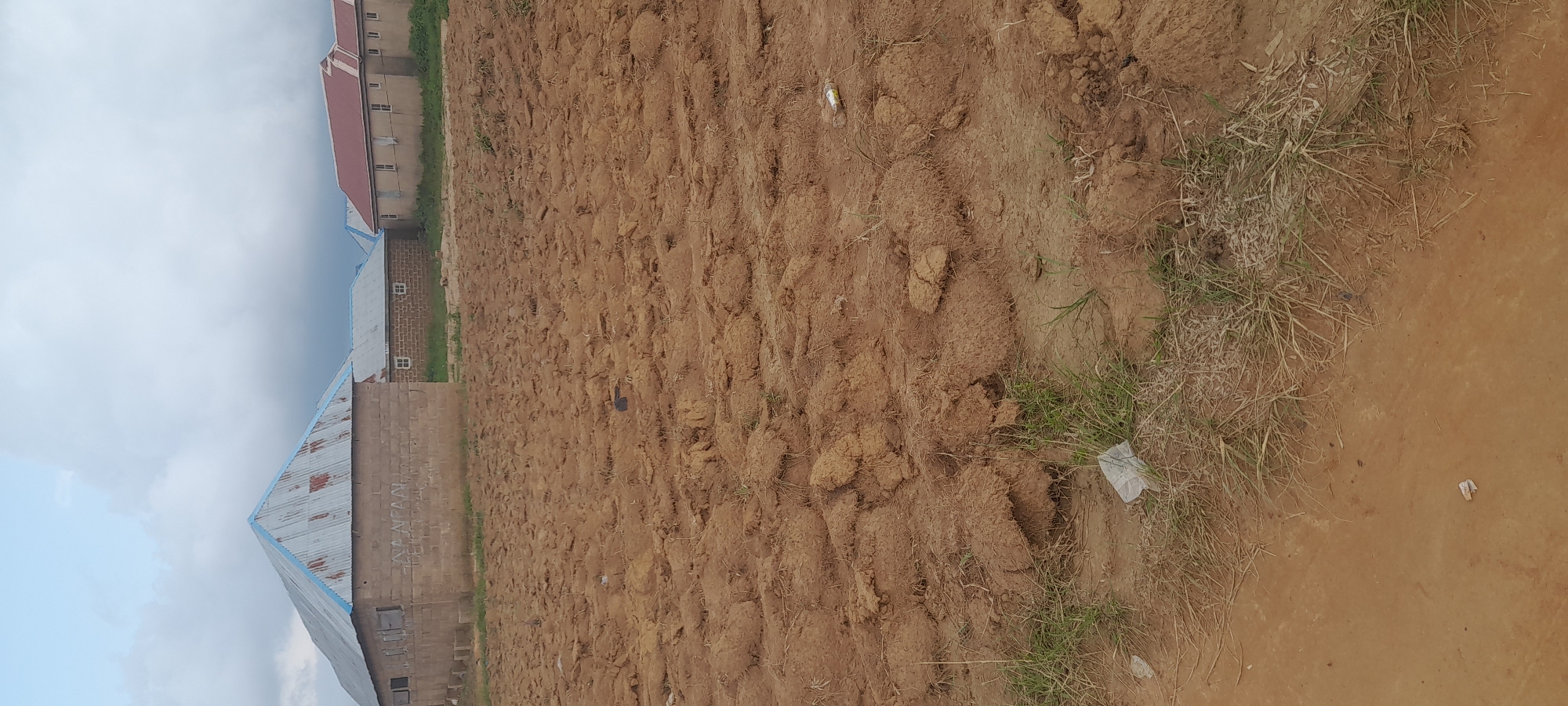 The image depicts a partially constructed or uncultivated plot of land with several buildings in the background. The foreground is dominated by rough, brown soil, indicating it has recently been tilled or disturbed. Clumps of earth are visible, suggesting uneven terrain. Sparse dry grass and some small pieces of white plastic litter are scattered across the soil.
In the background, three distinct buildings are visible. The leftmost building is a rectangular structure with a corrugated metal roof, painted with blue trim. Its walls appear to be constructed of concrete blocks. The middle building is smaller and also has a corrugated metal roof with blue trim. It has a visible window with a small square pane and a white border. To the right, a longer building with a red-tiled roof is situated. This building has multiple windows and appears more complete than the other two. The overall environment suggests a rural or developing suburban area, possibly on the outskirts of a city. The sky is overcast with significant cloud cover. The specific location of Durumi, Nigeria, is noted.