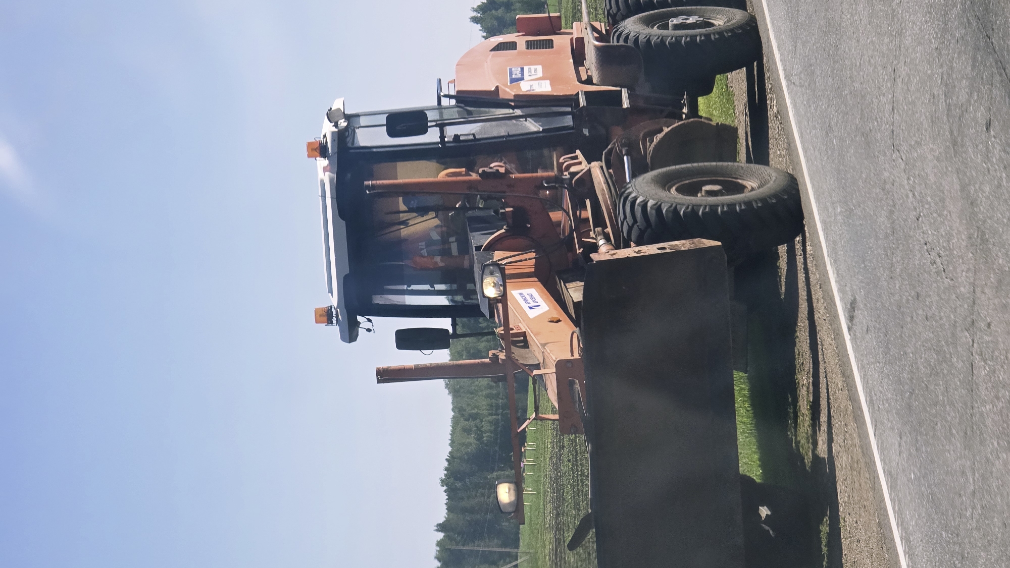 An orange road grader is parked on the side of an asphalt road. The grader is in the foreground, facing slightly to the right. It has a large metal blade at the front, large rubber tires, and a cab with windows. There are orange flashing lights on the roof of the cab. A person wearing an orange high-visibility vest is visible inside the cab. In the background, there is a green field with some vegetation and trees. The sky is clear and blue. The image appears to be taken during the daytime.