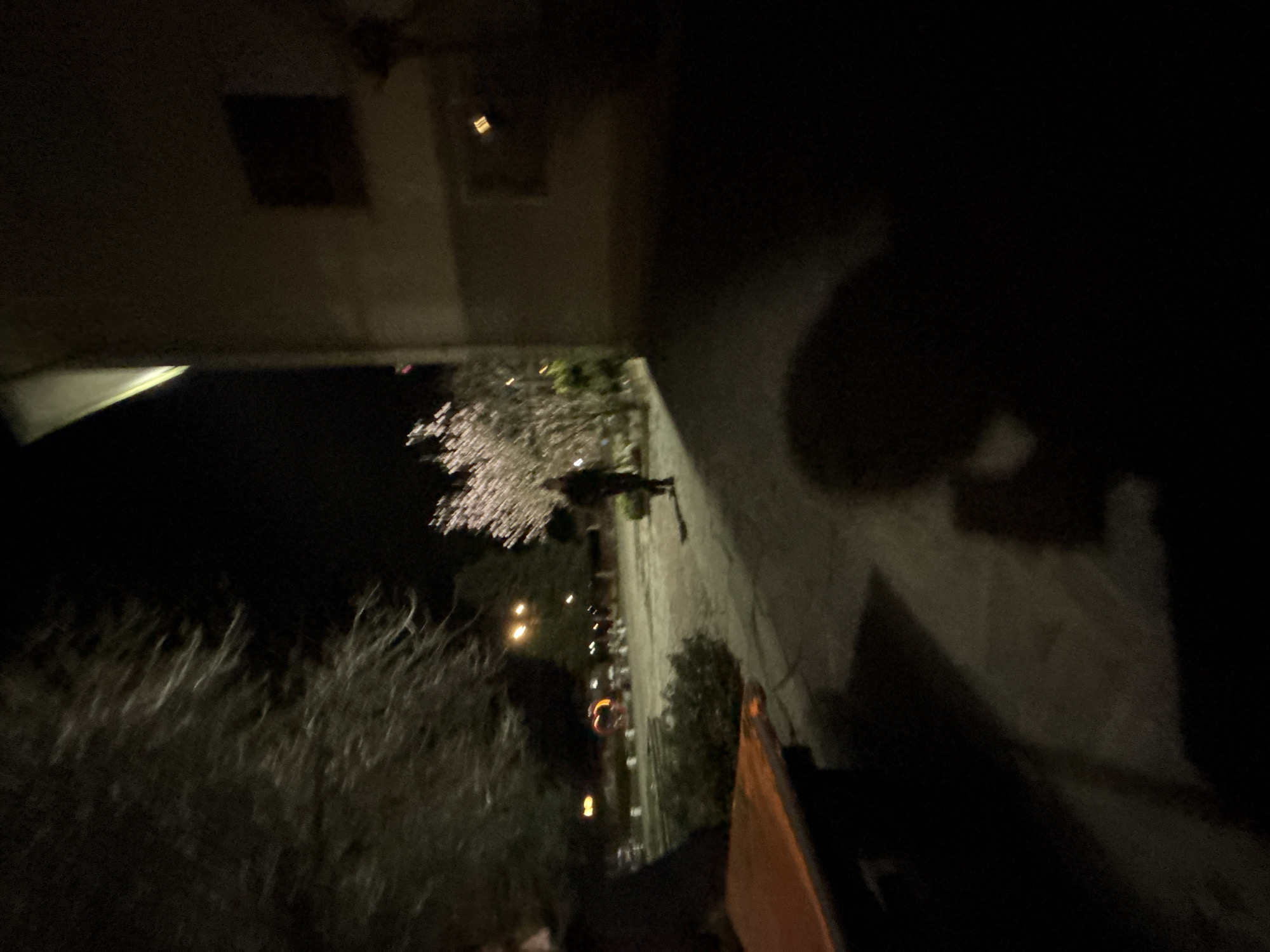 The image shows a nighttime scene, seemingly taken from an elevated position, perhaps a balcony. A building exterior is visible in the top and right portions of the frame. The lower portion shows trees, some with blossoming flowers illuminated by streetlights in the background. The photograph doesn't reveal enough detail to determine the precise location within Kliment, Bulgaria. There are no visible people or actions taking place in the image.