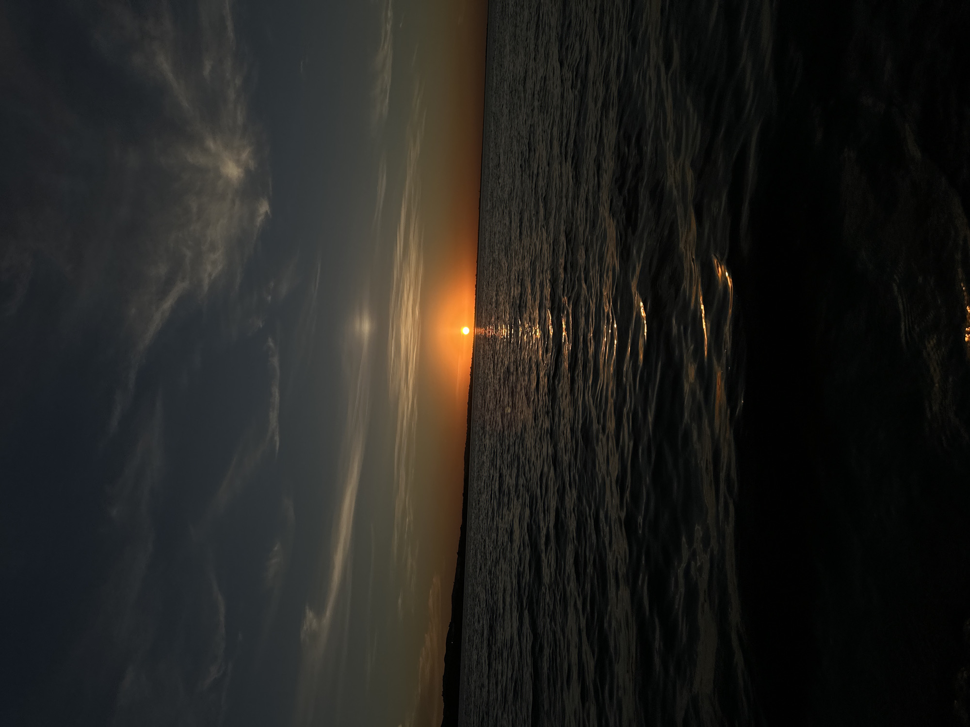 A wide-angle, low-angle shot captures a tranquil sunset over a body of water. The sun, a bright orange orb, is positioned low on the horizon, casting a shimmering golden path across the dark, choppy water. Wispy clouds with a subtle pinkish hue are scattered across the deep blue and grey sky. A dark, silhouetted landmass with gentle hills is visible in the distance along the left side of the horizon. The overall impression is one of peaceful solitude and natural beauty, likely observed from a boat or the water's edge in Kokshetau, Kazakhstan, a city known for its scenic lakes. There are no people or discernible objects in the image, focusing solely on the expansive natural landscape.
