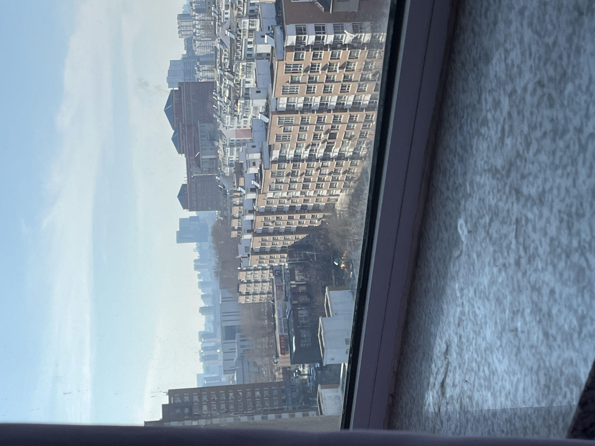 The image shows a view from a window in Changchun City, China. The cityscape is dominated by high-rise apartment buildings with light brown or white exteriors and rows of windows. In the distance, the sky is pale blue with some light clouds. The window frame is visible on the right side of the image, and the interior wall next to the window has a textured, uneven surface.
