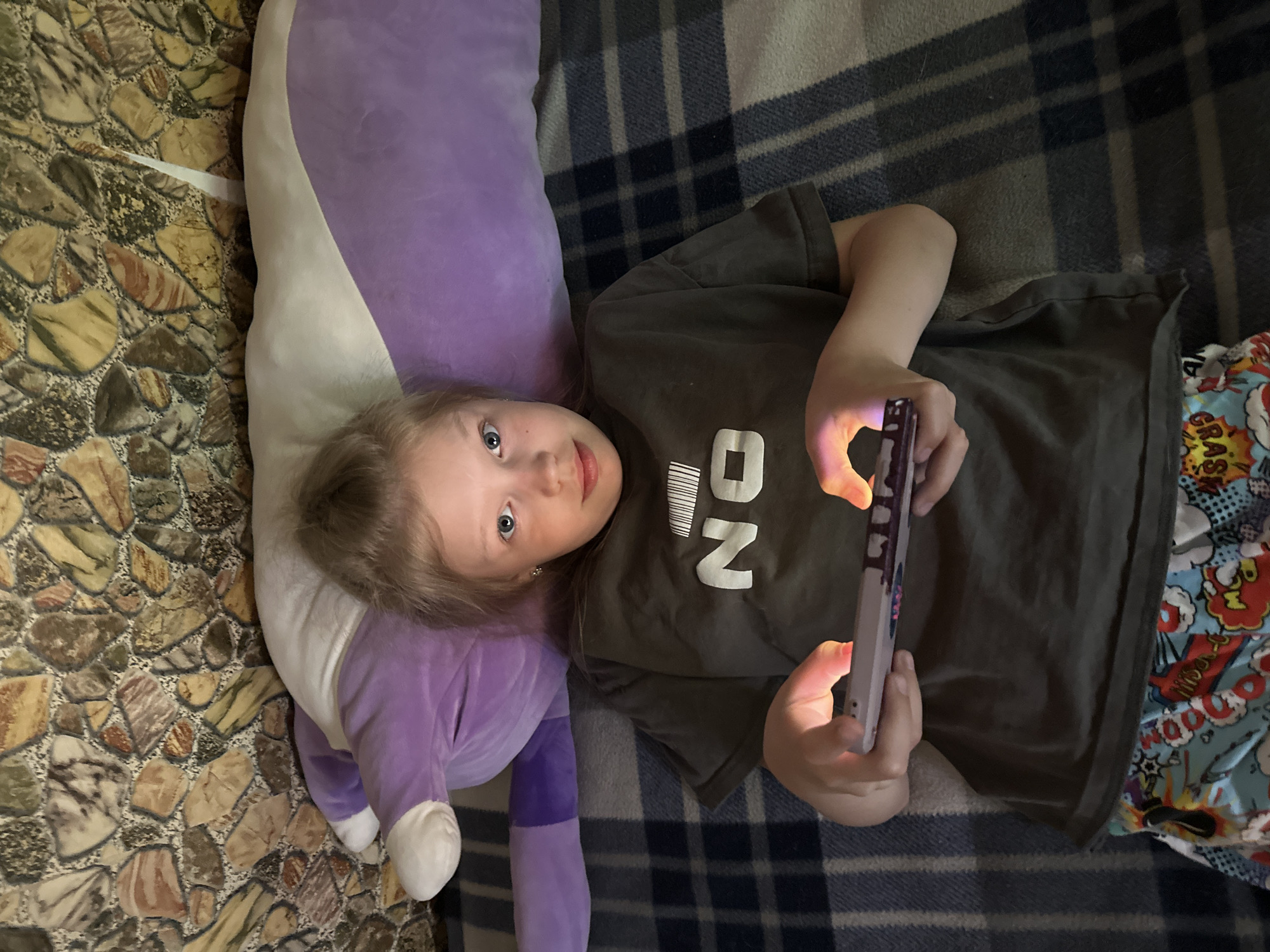 A young girl with blonde hair and blue eyes is lying on a plaid blanket, propped up against a large purple pillow. She is wearing a dark gray t-shirt with the word "NO" printed on it in white block letters, and colorful pajama bottoms adorned with comic book graphics. Her hands are holding a tablet, which is illuminated by its screen, casting a soft glow on her fingers. The background appears to be a textured wall with a stone-like pattern. The scene suggests a relaxed, indoor setting, possibly a bedroom or living room, in a residential area of Sayanogorsk, Russia.