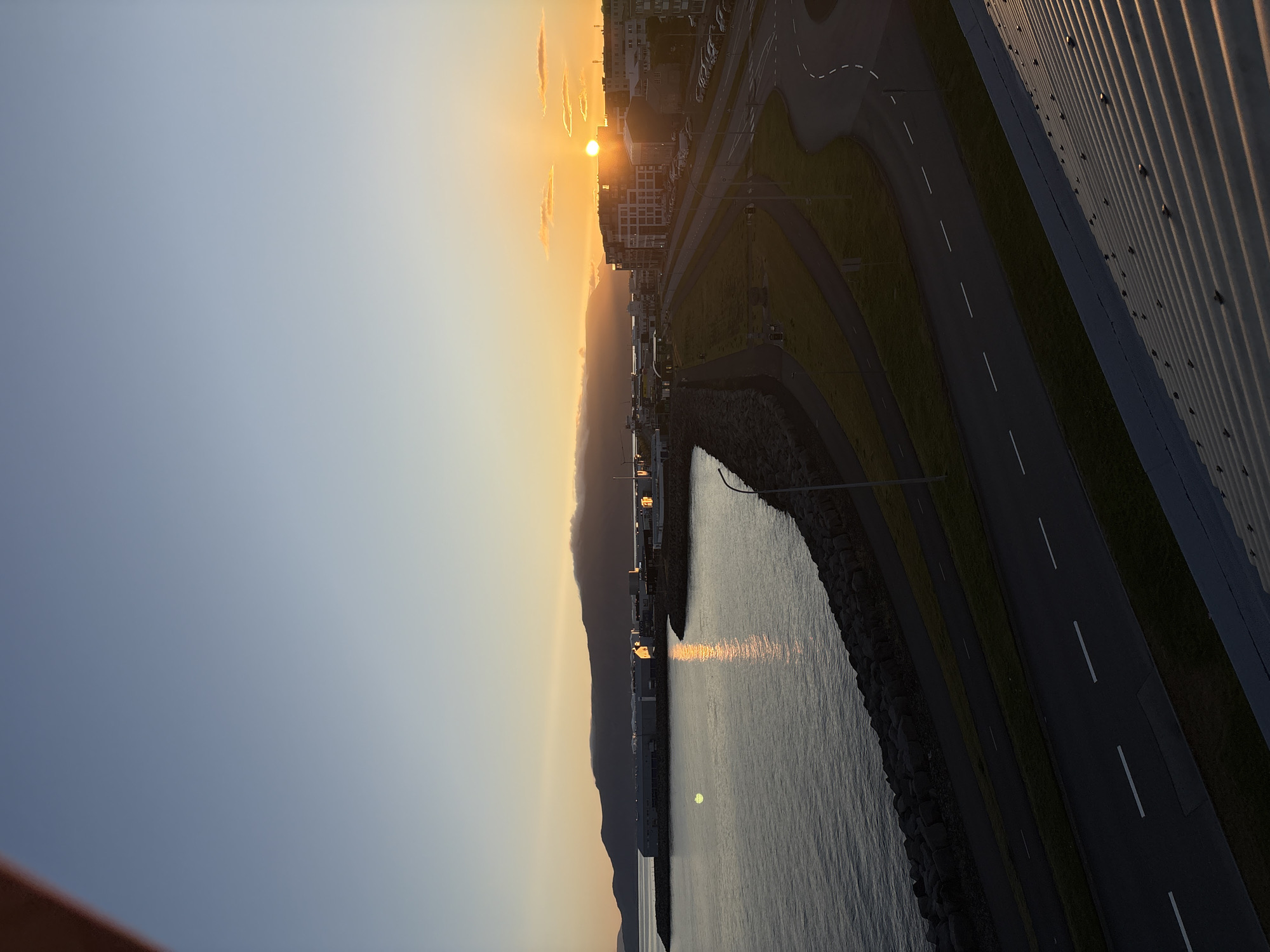 This image captures a panoramic view of Reykjavik, Iceland, during what appears to be a summer sunset. The foreground is dominated by a corrugated metal roof, casting a shadow over the lower portion of the frame. To the left, a dark, rocky shoreline separates the choppy, dark blue water of the ocean from a paved path and a grassy embankment. Several modern, low-rise buildings with flat roofs line the waterfront, their windows reflecting the warm light of the setting sun. Further inland, a multi-story building with a contemporary facade stands out amongst other structures. A network of roads, marked with white lines, winds through the scene, with parked cars visible along one of the streets. In the distance, a range of dark, mist-shrouded mountains forms a dramatic backdrop, their peaks softened by the low light. The sky transitions from a clear, pale blue at the top to a warm orange and yellow hue near the horizon, where the sun, a brilliant orb, is partially obscured by wispy clouds. The overall impression is one of a serene, urban coastal landscape bathed in the ethereal glow of the Arctic summer twilight.