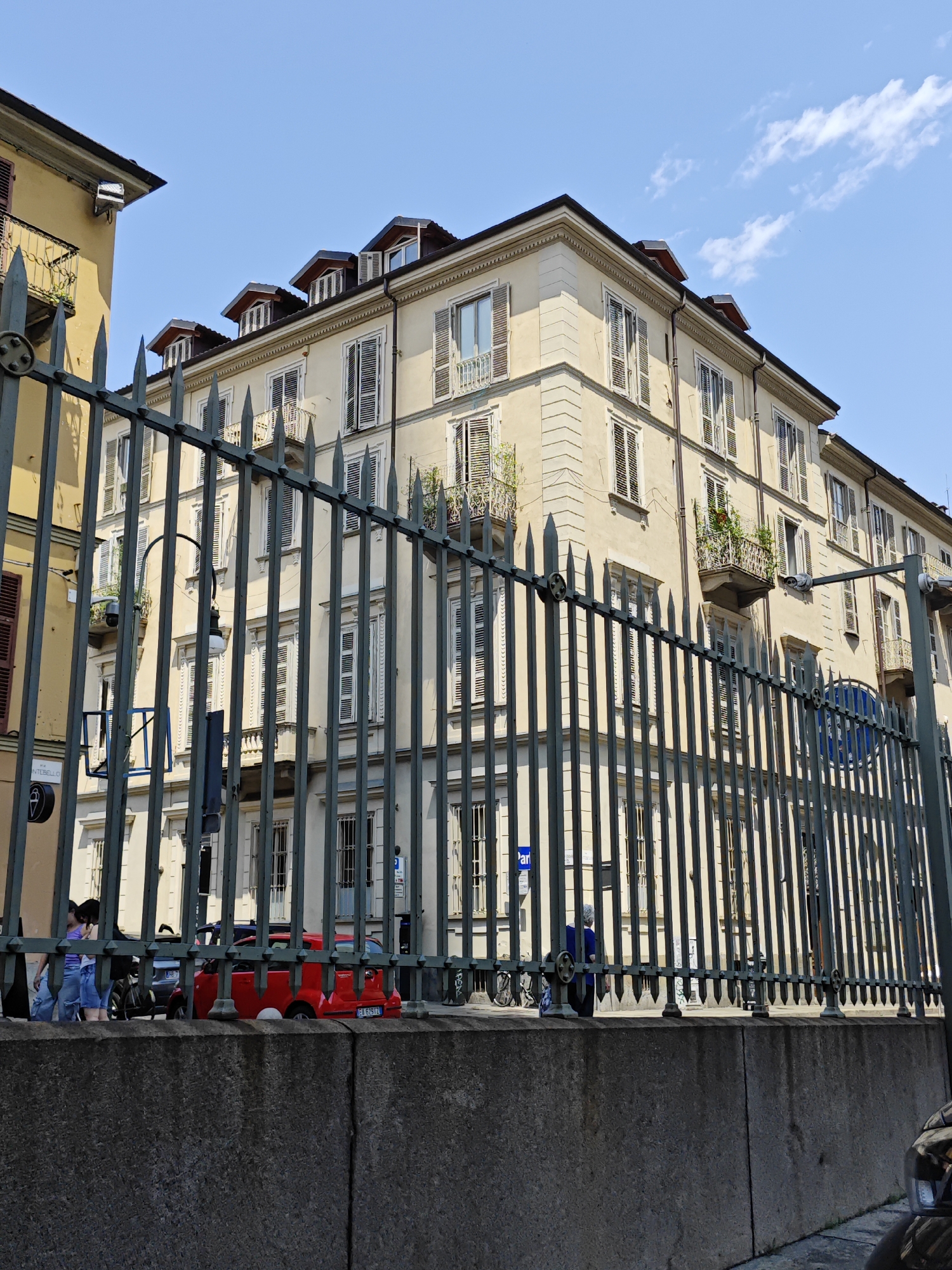 The image shows a light yellow building with multiple stories and many windows with closed wooden shutters. The building has a classical architectural style, with decorative elements such as cornices and balconies. In the foreground, there is a dark metal fence. Behind the fence, on the sidewalk, there are a few people, some cars, and a street lamp. The sky is blue with a few white clouds. The scene appears to be a street in Turin, Italy, specifically on Via San Donato near the intersection with Via Montebello. The time of day is likely late morning or early afternoon, given the bright sunlight.