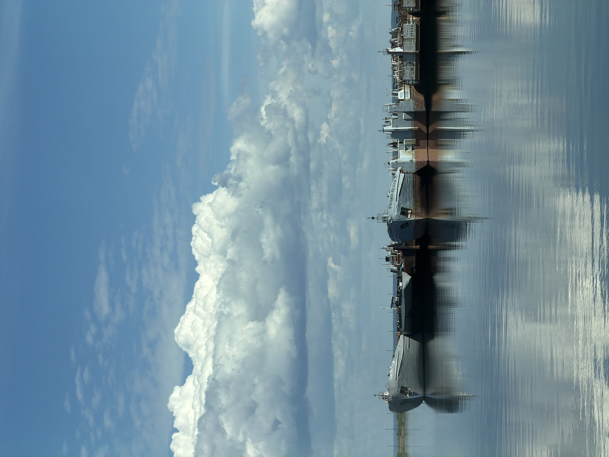 The image displays a static daytime scene featuring a line of industrial vessels moored on a calm body of water under a partially cloudy sky in Chaikovsky, Russia.
The upper approximately one-third of the frame is dominated by a clear, light blue sky transitioning to a band of dense, white cumulus clouds with some grey undersides extending across the middle third of the image. The clouds are predominantly layered horizontally.
The lower two-thirds of the image depict a wide, calm body of water, likely a river or reservoir, reflecting the sky and the vessels. The water surface exhibits subtle ripples, causing slight distortion in the reflections.
In the middle ground, horizontally aligned, a series of large, grey and rust-toned industrial vessels are closely moored. These vessels appear to be barges or cargo ships, characterized by flat decks and various superstructures, cabins, and rigging details. The leftmost vessel prominently displays Cyrillic lettering on its hull, reading "ПРИСТАВКА-ВОЛЖСКИЙ" (Pristavka-Volzhsky), and another vessel shows the number "3105." The vessels are reflected with moderate clarity in the water below. Behind the vessels, a narrow strip of distant land or shoreline is visible, sparsely vegetated, with faint vertical structures possibly indicating power lines or markers. No human figures or active movement are present within the scene. The overall impression is one of an inactive industrial waterfront.