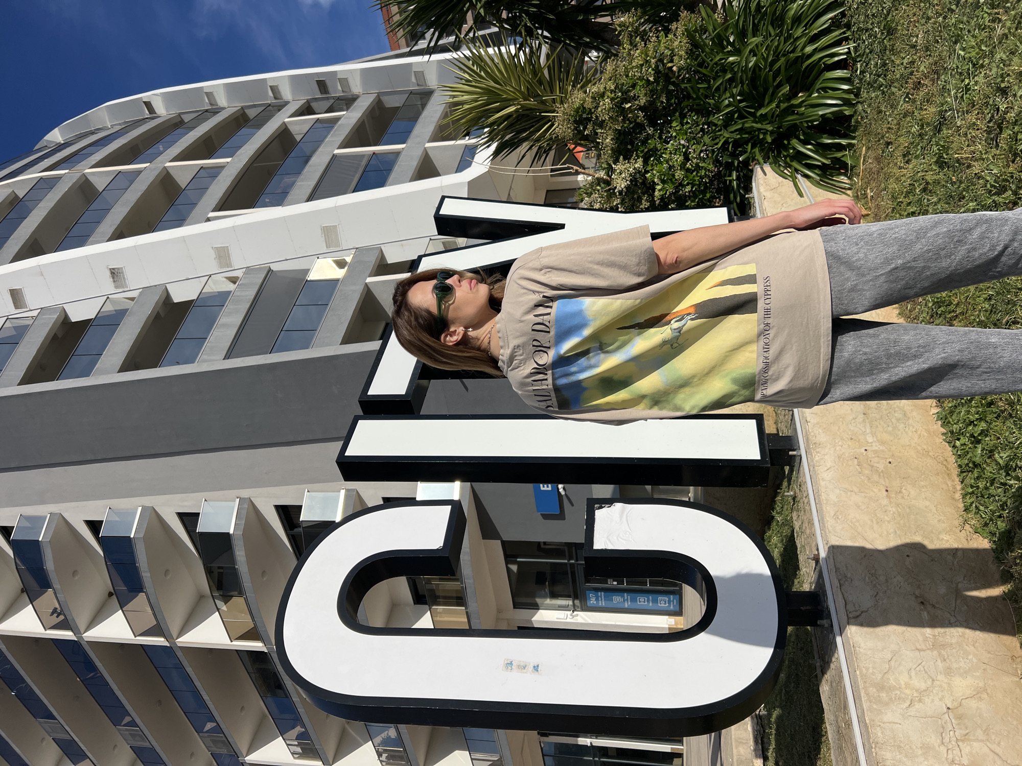 A medium-shot photograph captures a woman posing with the word "VI" in front of a multi-story building. The woman, wearing sunglasses and a tan t-shirt with grey sweatpants, is centered in the foreground. The large, white, and black "VI" sign dominates the middle ground. In the background stands a tall building with a modern architectural design that features repeating geometric shapes. Palm trees and greenery are also in the background. The picture appears to be taken during the day under clear, sunny skies. Based on the sign and architectural features, the location is likely near the Swisslion Hotel Vile Oliva in Bar, Montenegro.