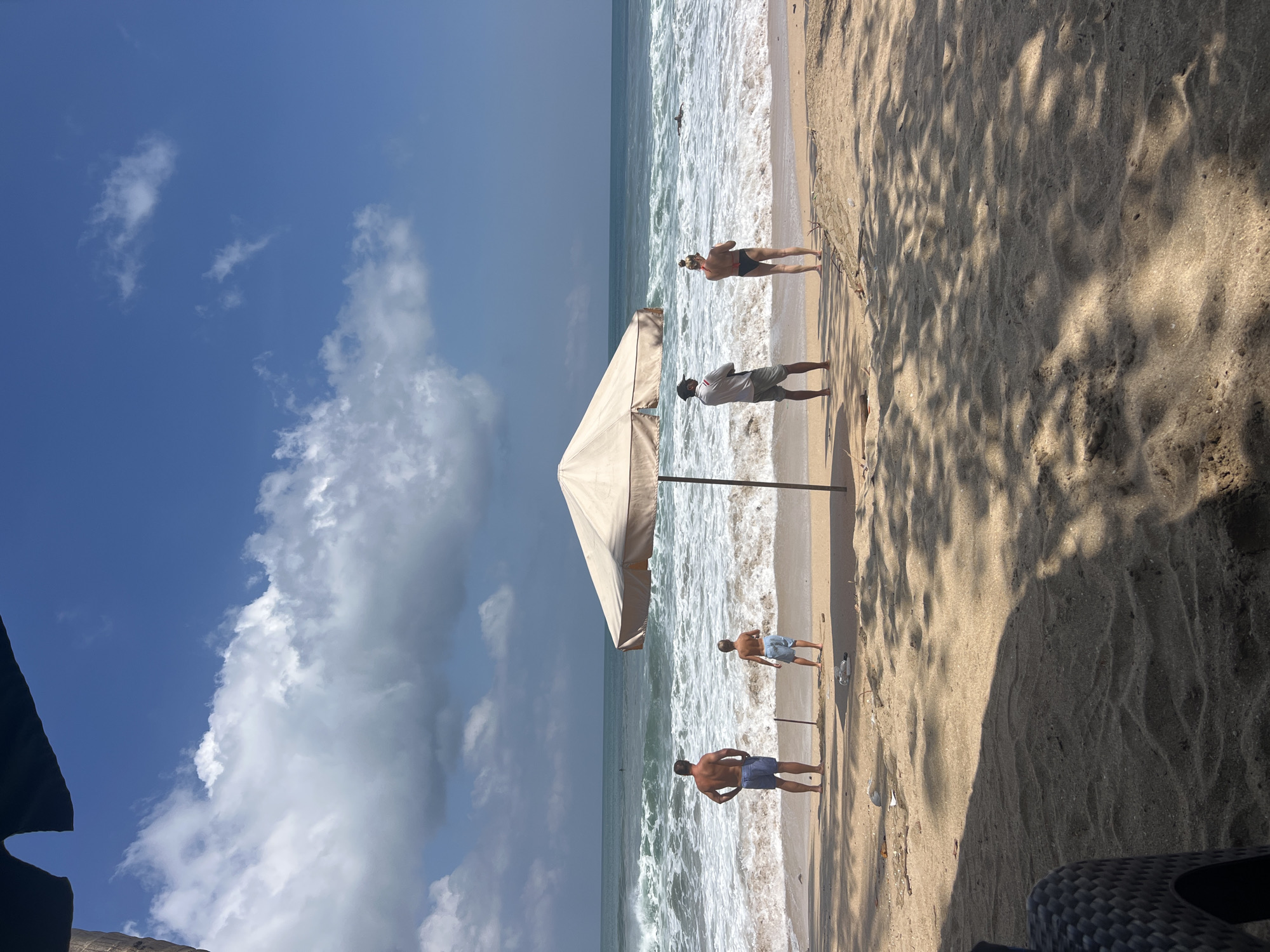 The media file depicts a beach scene in Kuta, Indonesia. The image shows several individuals enjoying a day at the beach. In the foreground, a large beach umbrella casts a shadow over the sand. Two men are visible, one standing closer to the ocean and another further back, both appearing to be relaxed or observing the surroundings. Further up the beach, another man is partially visible, and a woman in swimwear is seen standing near the water.
The ocean is active with waves breaking on the shore, suggesting moderate to rough conditions. The sky is a mix of blue with scattered white clouds, indicating a bright and possibly warm day. The overall setting is a typical tropical beach with sand, sea, and sky. The presence of the umbrella suggests a desire for shade from the sun. The people are dressed in beach attire, implying a leisurely activity such as sunbathing, swimming, or simply enjoying the coastal environment. The image captures a moment of leisure and relaxation in a popular tourist destination. There is no visible text in the media file.