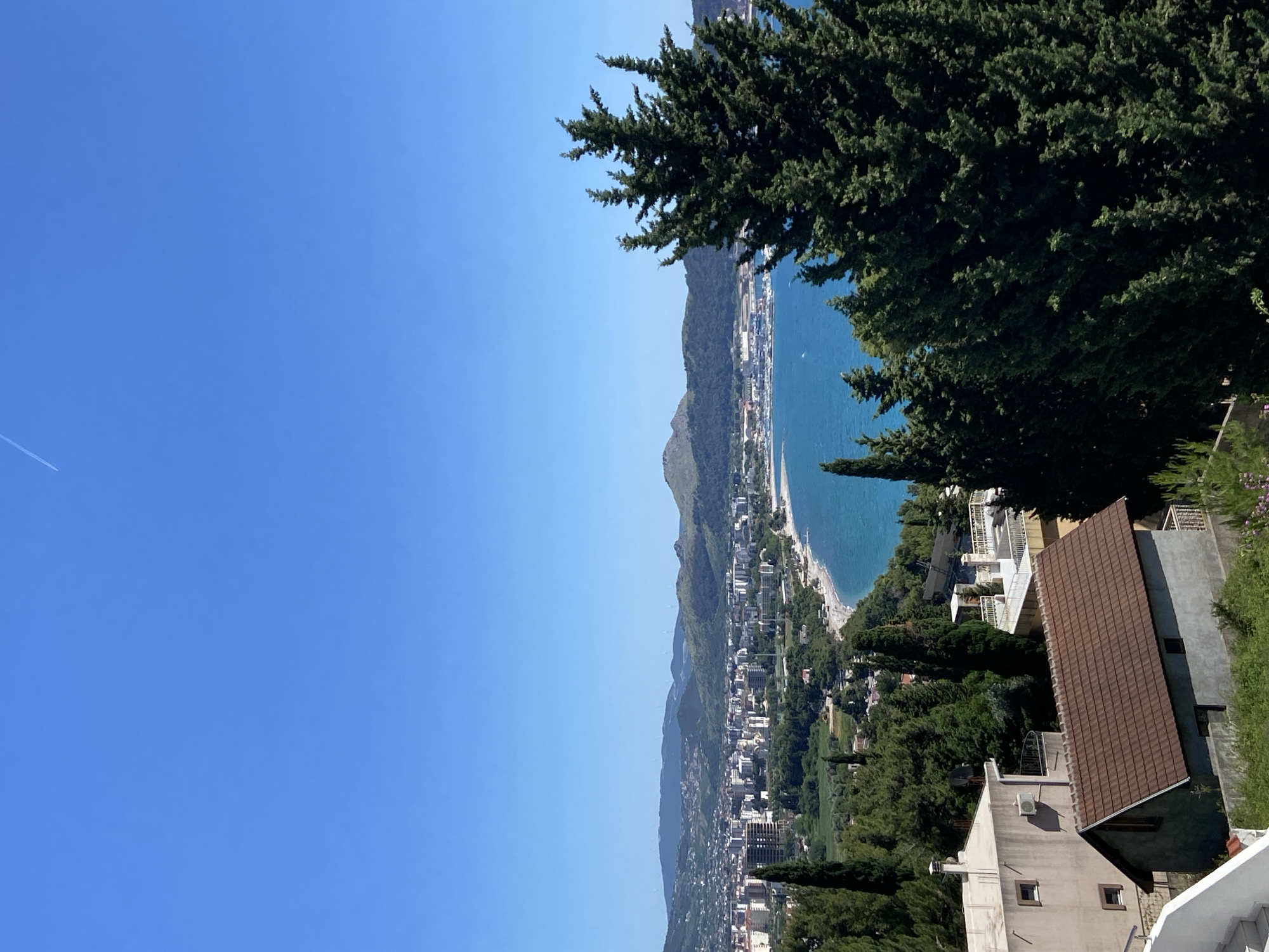 The image shows a coastal cityscape, specifically Šušanj, Montenegro. The scene is oriented vertically. To the left, a clear blue sky takes up a significant portion of the frame, with a contrail faintly visible. A coastal city stretches along the base of mountains; buildings, vegetation, and a beach are distinguishable. In the foreground on the right, parts of several buildings are visible, including one with a brown tiled roof. A large conifer tree dominates the upper right corner of the image.