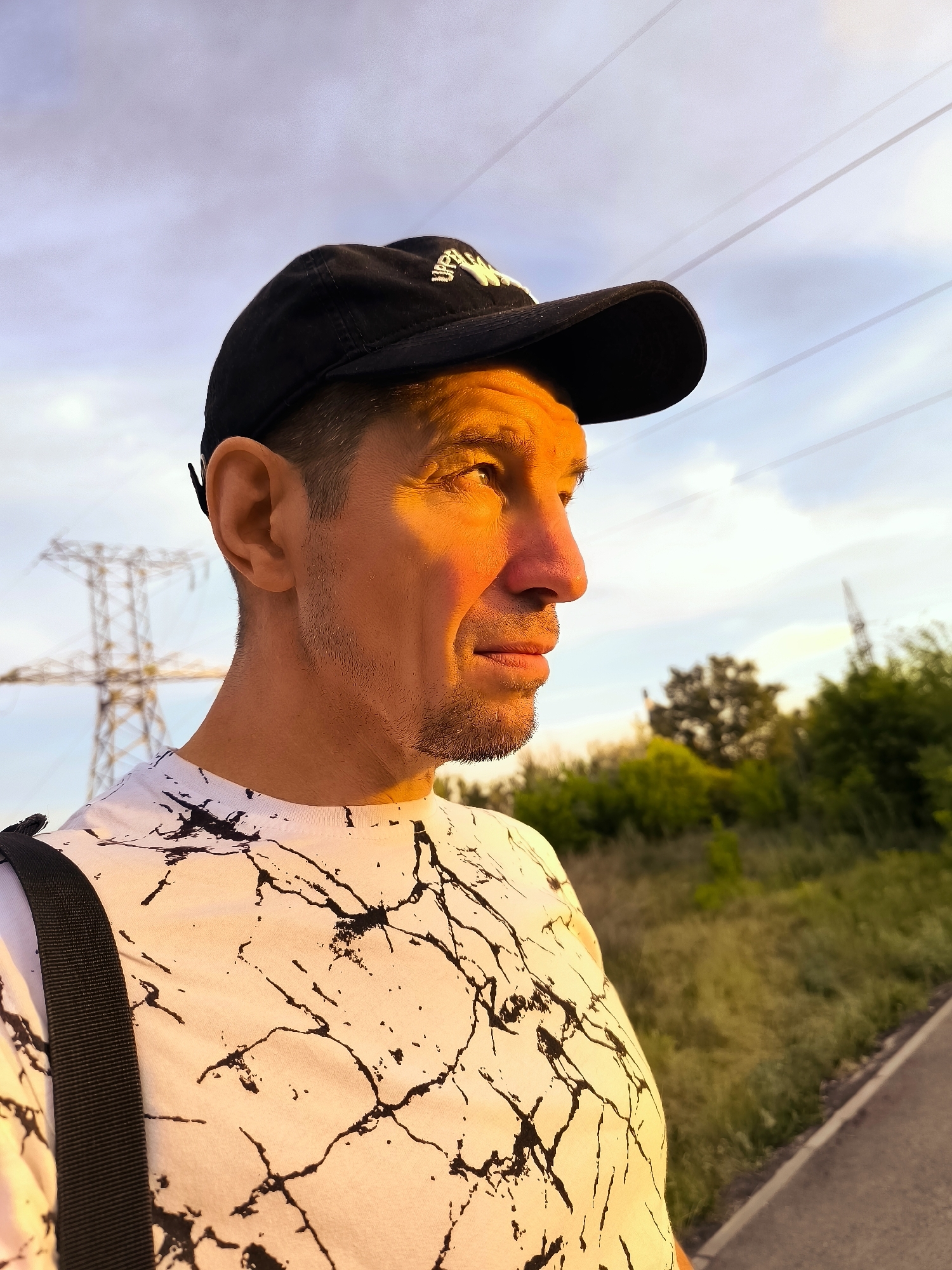 In a close-up, side-profile shot, a man wearing a black baseball cap and a white t-shirt with a black marble-like pattern is illuminated by the warm, golden light of a sunset. The light casts a strong orange hue across his face, highlighting his features and creating a dramatic contrast with the shadowed side of his face. He is looking off to the right of the frame. In the background, a blurry landscape of green foliage and a tall, skeletal power line tower are visible against a pale sky with thin, horizontal clouds. The scene appears to be outdoors, possibly on the outskirts of a city, given the presence of the power lines and tower.