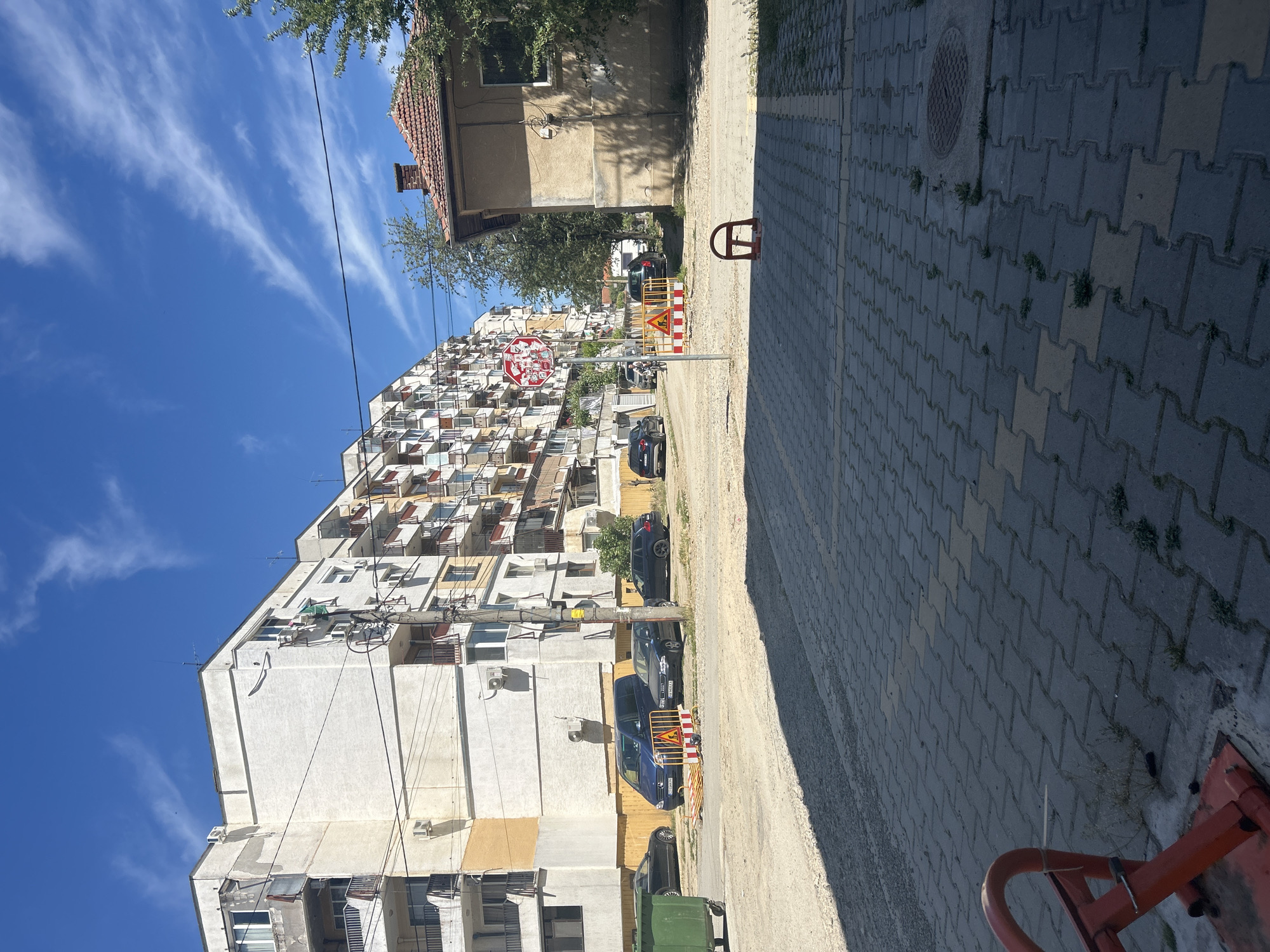 The image depicts a street scene in Kazanlak, Bulgaria. An apartment building with multiple balconies faces the street. Several parked cars are visible along the building's side. A stop sign and roadwork barriers are present, suggesting construction or maintenance work is in progress. Across the street, a small house with a red tile roof stands next to a sidewalk made of interlocking paving stones. The sky is blue with scattered clouds. There are no people visible in the image.
