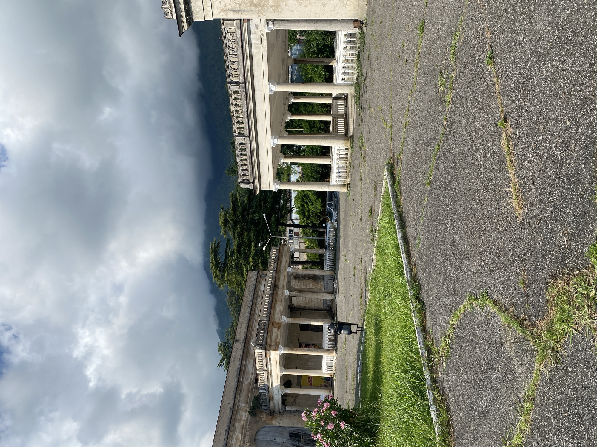 This is an eye-level, outdoor, full shot of a dilapidated building with columns and a courtyard in Psakhara, Abkhazia. The building, constructed of aged concrete, has a covered walkway supported by numerous white columns. Above the walkway is a decorative railing, also white, with intricate carvings. A pale, weathered facade runs along the top of the structure. In front of the building, a paved courtyard shows signs of disrepair, with cracks and weeds growing through the asphalt. To the left, a patch of vibrant green grass is bordered by a concrete curb. A lone black trash receptacle stands in the courtyard. Behind the building, lush green trees and mountains shrouded in a dramatic, cloudy sky create a backdrop. The overall scene suggests an abandoned or neglected historical site.