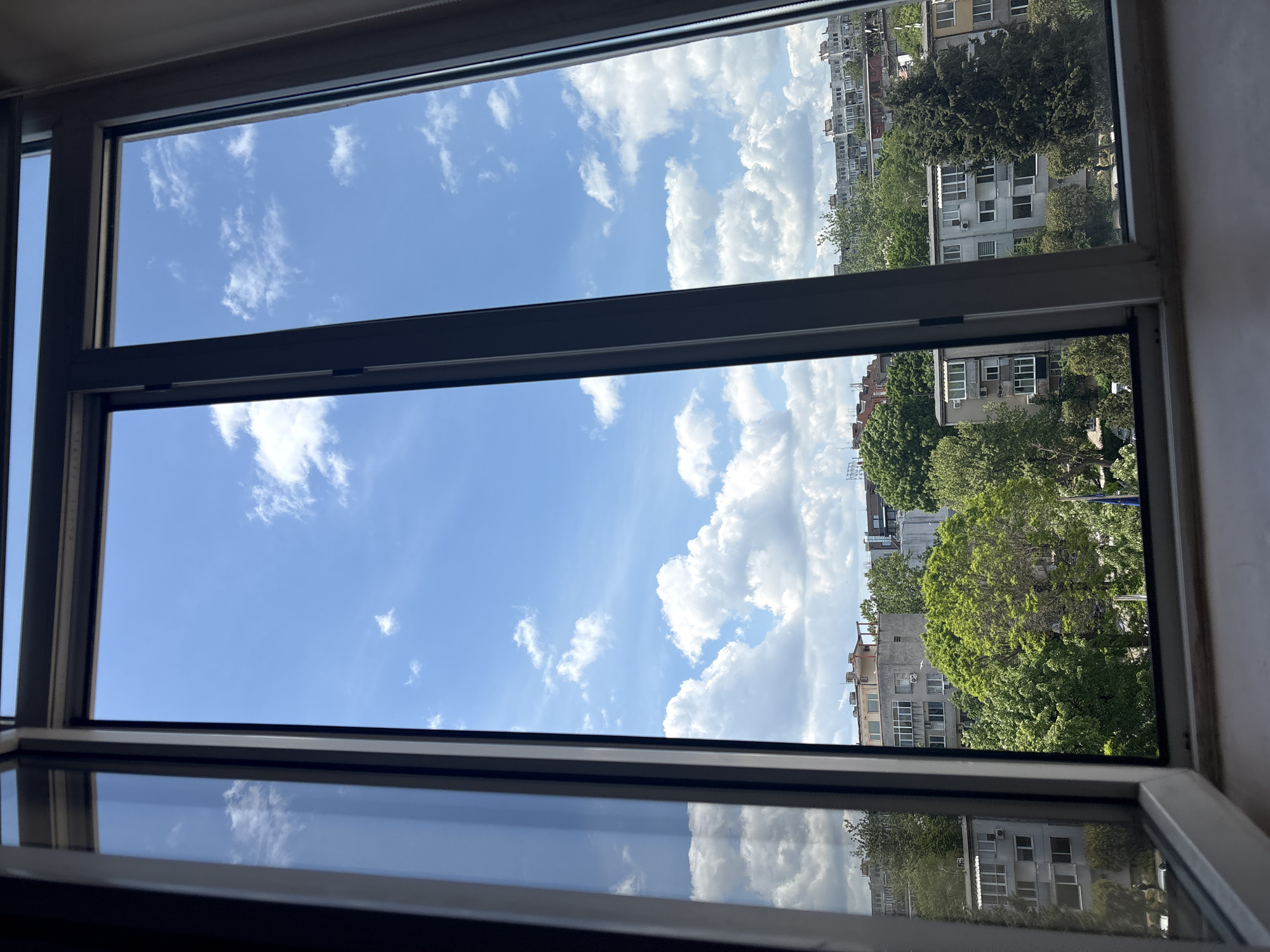 The image depicts a view from an open window. The sky is blue with scattered white clouds. Outside the window, there are buildings and trees, indicating an urban setting. The location is the city of Burgas, Bulgaria. The window frames are visible, and the sky's reflection is seen on the open lower part of the window.