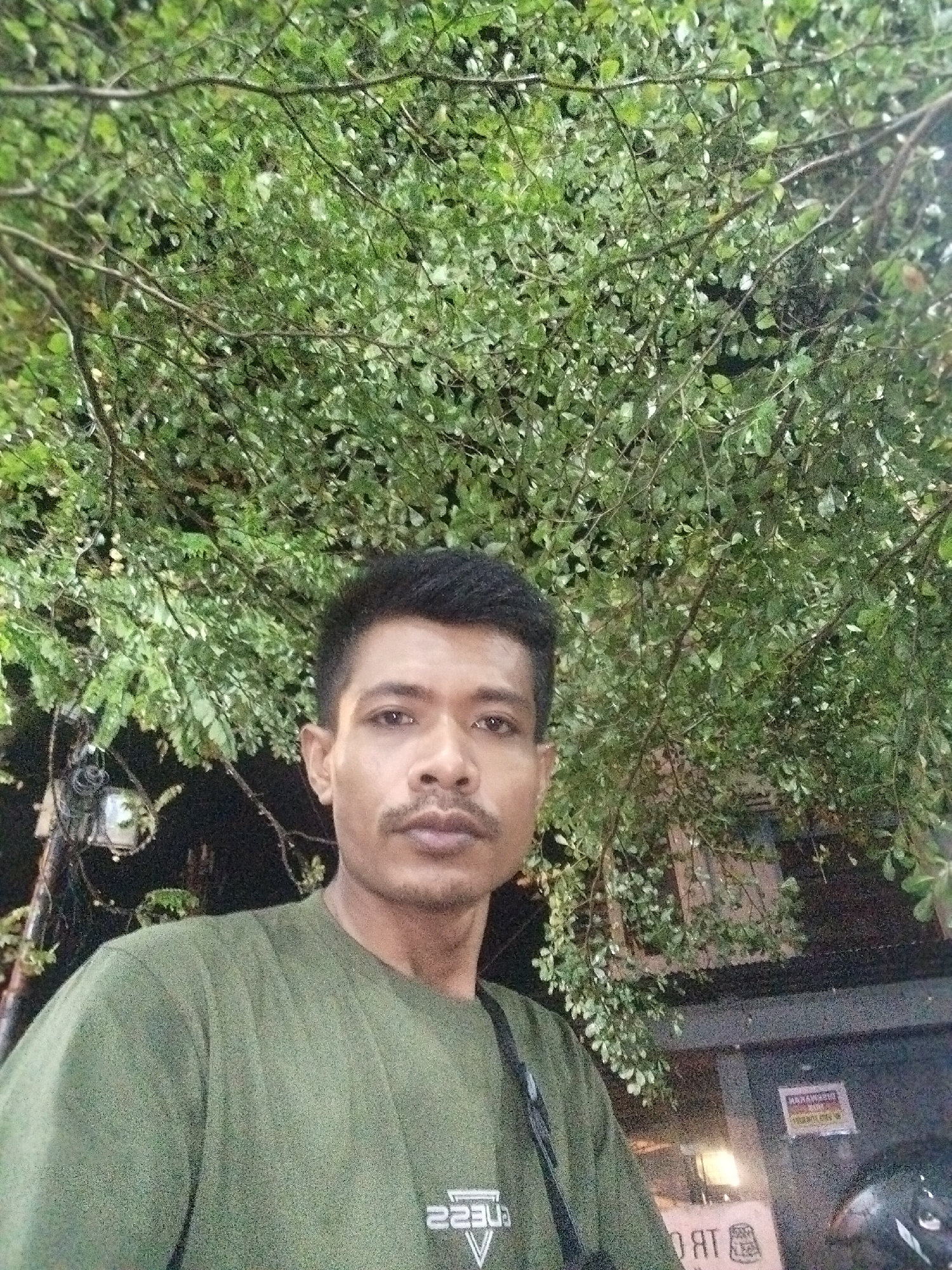 A person stands outdoors under a tree with dense green foliage. The background is dark, suggesting it is night. A street light is visible to the left, and a building is seen behind the person with a sign. A black motorcycle helmet is also present. The image may have been taken in Lhokseumawe, Indonesia.