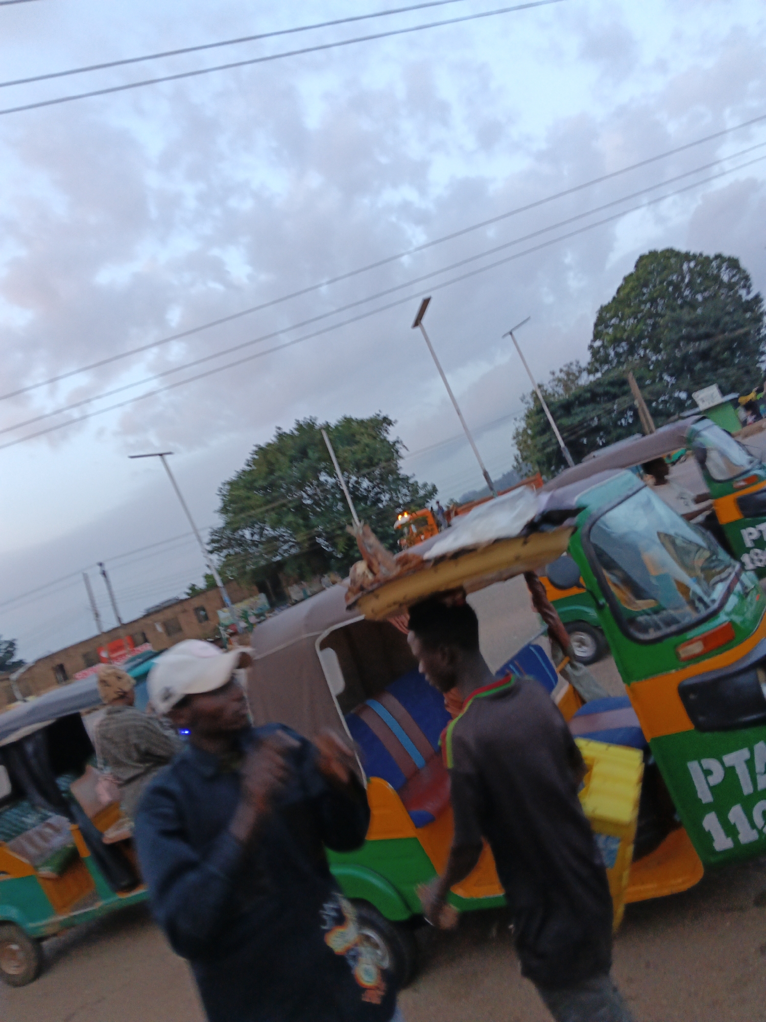 In Bukuru, Nigeria, an overcast sky with scattered clouds hangs above a street scene with several auto-rickshaws, also known as Keke Napep. Two men are the central focus. In the foreground, a man wearing a white baseball cap and a dark blue long-sleeved shirt is facing away from the camera, his arms in motion as if gesturing or in the process of being captured in action. To his right, another man in a dark grey t-shirt is walking away from the camera, carrying a bright yellow object. Behind them, the colorful auto-rickshaws are parked, one of which has "PTA 110" visible on its side. The auto-rickshaws have green and orange bodies and are equipped with seats and canopies. In the background, buildings and trees are visible, contributing to the urban and somewhat hazy atmosphere. The ground appears to be a dirt or unpaved road.