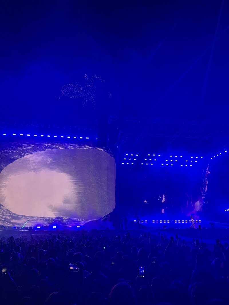 The image shows a large concert stage illuminated with blue lighting, filled with a crowd of people, likely taken in Indio, United States. A large screen dominates the left side of the stage, displaying imagery or content in a light color, while the right side is filled with smaller screens and stage lighting, creating a vibrant and dynamic visual display. The crowd is dense, with many individuals holding up their phones to record the event. Above the stage and crowd, in the blue lighting, a silhouette of a figure, potentially formed by drones, is visible.