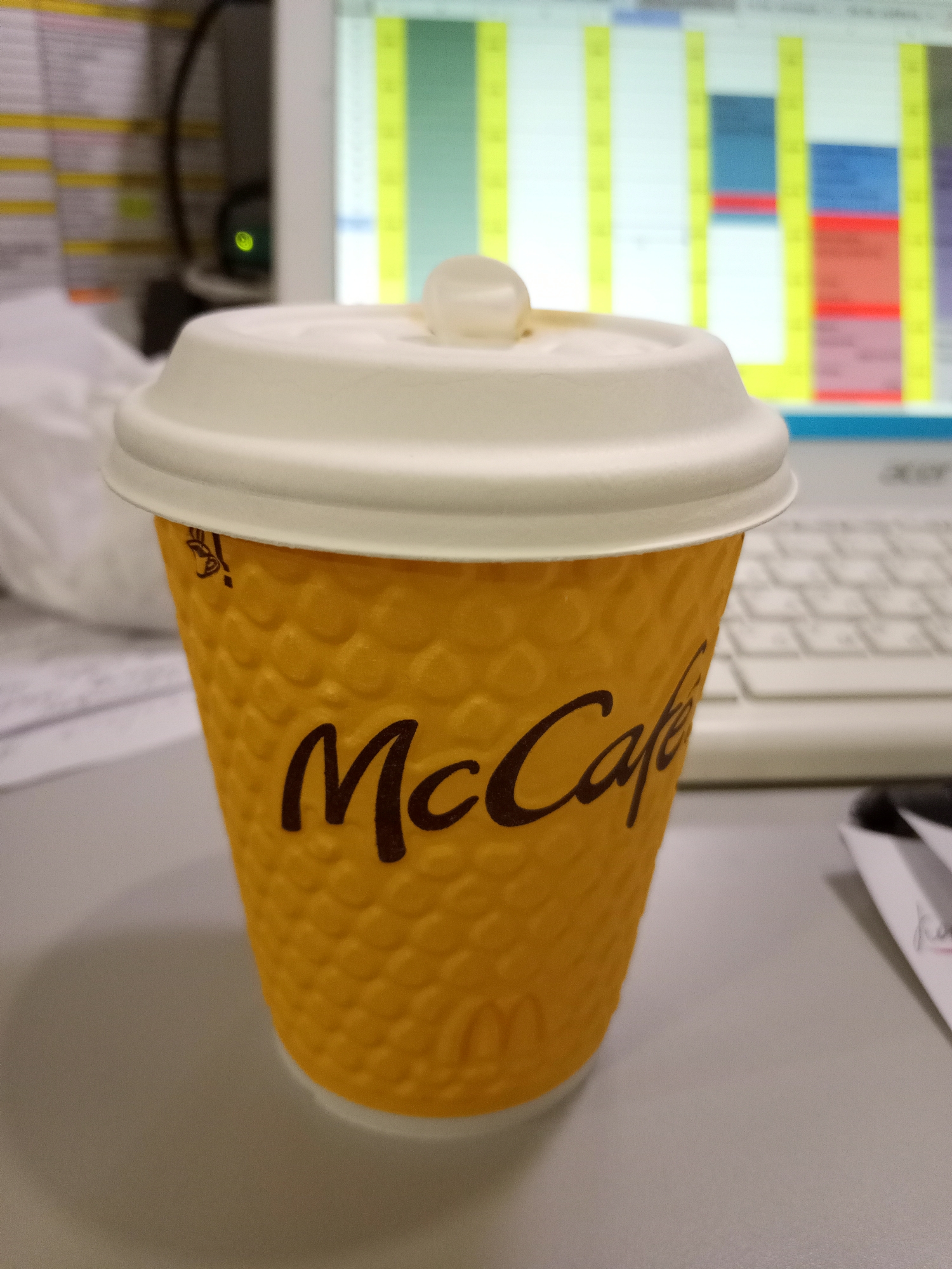 A close-up, eye-level shot shows a McCafe paper cup with a white lid. The cup is bright yellow with a textured pattern and the McCafe logo printed in dark brown. The background is blurred, showing a computer monitor displaying a colorful calendar or schedule, and a white keyboard. The overall setting appears to be an office environment.