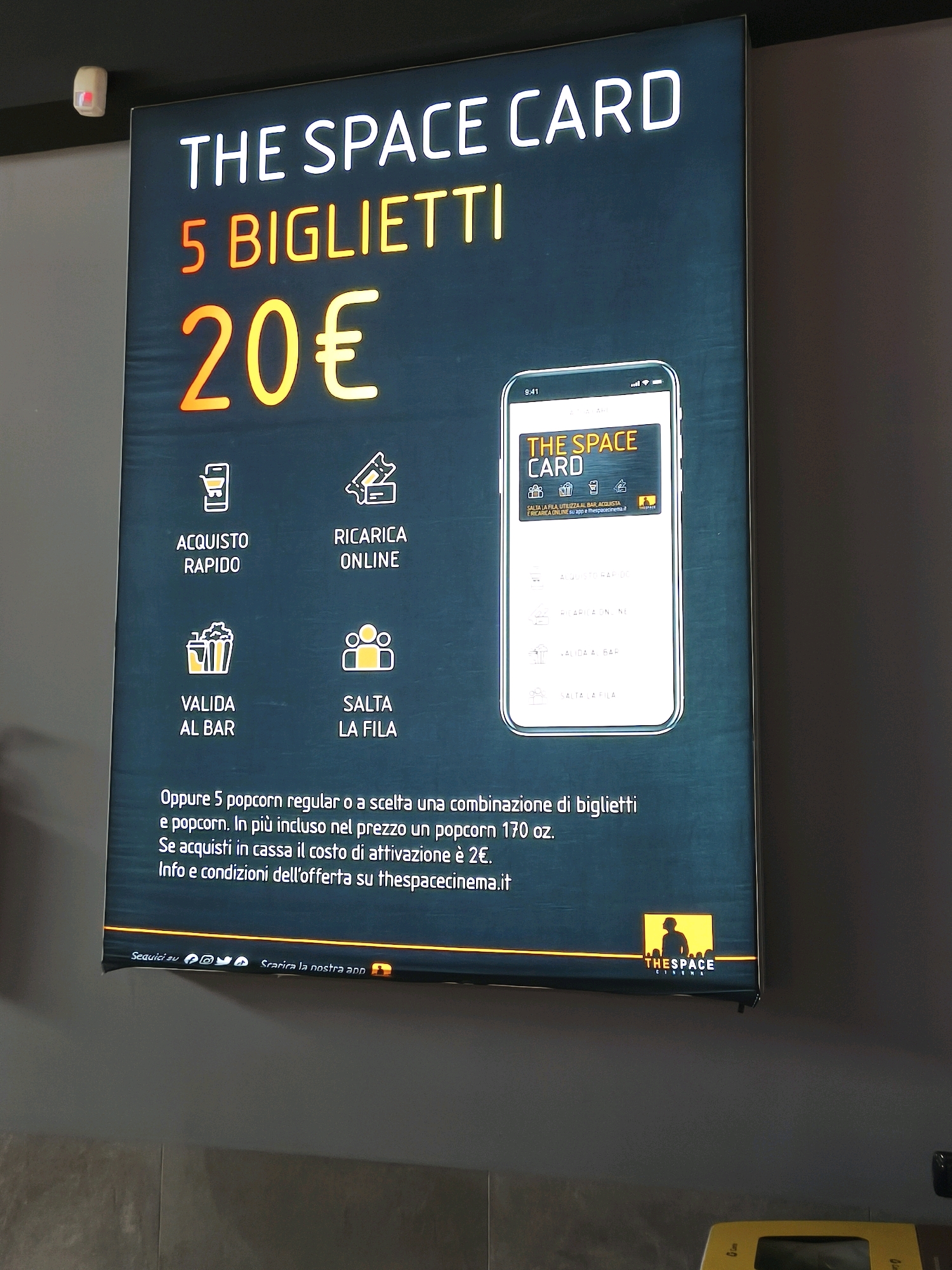 The image shows a large illuminated advertisement for "THE SPACE CARD" at a cinema. The advertisement displays the offer "5 BIGLIETTI 20€" in large, bold, neon-like lettering. Below this, there are four icons with accompanying text: "ACQUISTO RAPIDO" (Quick Purchase), "RICARICA ONLINE" (Online Recharge), "VALIDA AL BAR" (Valid at the Bar), and "SALTA LA FILA" (Skip the Line).
A smartphone screen is depicted within the advertisement, showing a digital representation of the "THE SPACE CARD" and its features.
Further text on the advertisement reads: "Oppure 5 popcorn regular o a scelta una combinazione di biglietti e popcorn. In più incluso nel prezzo un popcorn 170 oz. Se acquisti in cassa il costo di attivazione è 2€. Info e condizioni dell'offerta su thespacecinema.it". This translates to: "Or 5 regular popcorn or a combination of tickets and popcorn. Also included in the price is a 170 oz popcorn. If you purchase at the counter, the activation cost is €2. Offer info and conditions at thespacecinema.it".
At the bottom of the advertisement, social media icons and the text "Scarica la nostra app" (Download our app) are visible, alongside the logo for "THE SPACE CINEMA".
The advertisement is mounted on a dark wall, likely within a cinema lobby. The lighting is primarily from the advertisement itself, casting a glow on the surroundings. There are no people clearly visible in the frame, nor any specific indicators of the city of Chiugiana, Italy. The scene is static and appears to be a promotional display.