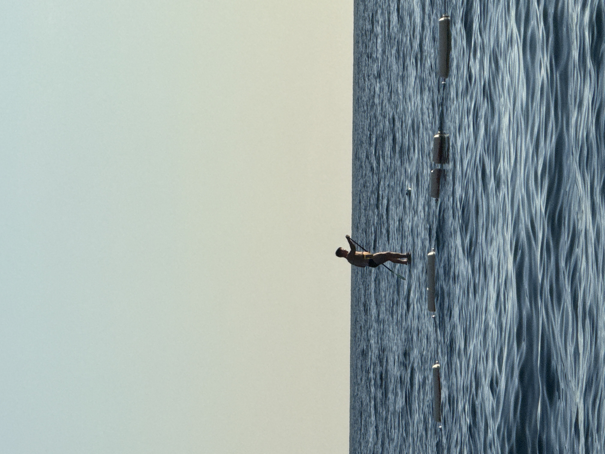 In Pržno, Montenegro, a lone individual stands on a paddleboard in the calm, blue sea. The person, silhouetted against the pale sky, holds a paddle, appearing to be in motion. Several cylindrical buoys are visible in the water, some in a line in the foreground and others further out. The horizon is clearly defined, separating the sea from the soft gradient of the sky.