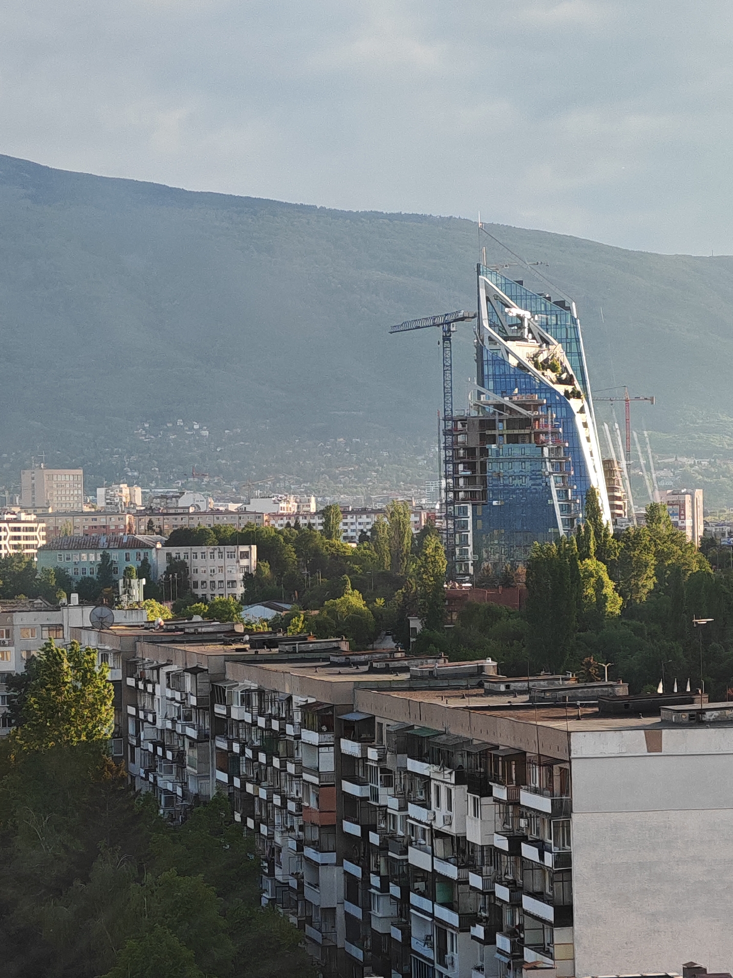 This is an eye-level shot taken in the city of Sofia, Bulgaria. The image focuses on an under-construction skyscraper in the city's skyline. The skyscraper is made primarily of glass and features a curved design. Cranes are visible around the skyscraper, indicating ongoing construction. Behind the skyscraper, a large mountain fills the horizon. In the foreground, there are dense, Soviet-era apartment blocks, characterized by their uniform architecture and multiple balconies. Green trees fill the spaces between the buildings. The sky is overcast with a slight haze.