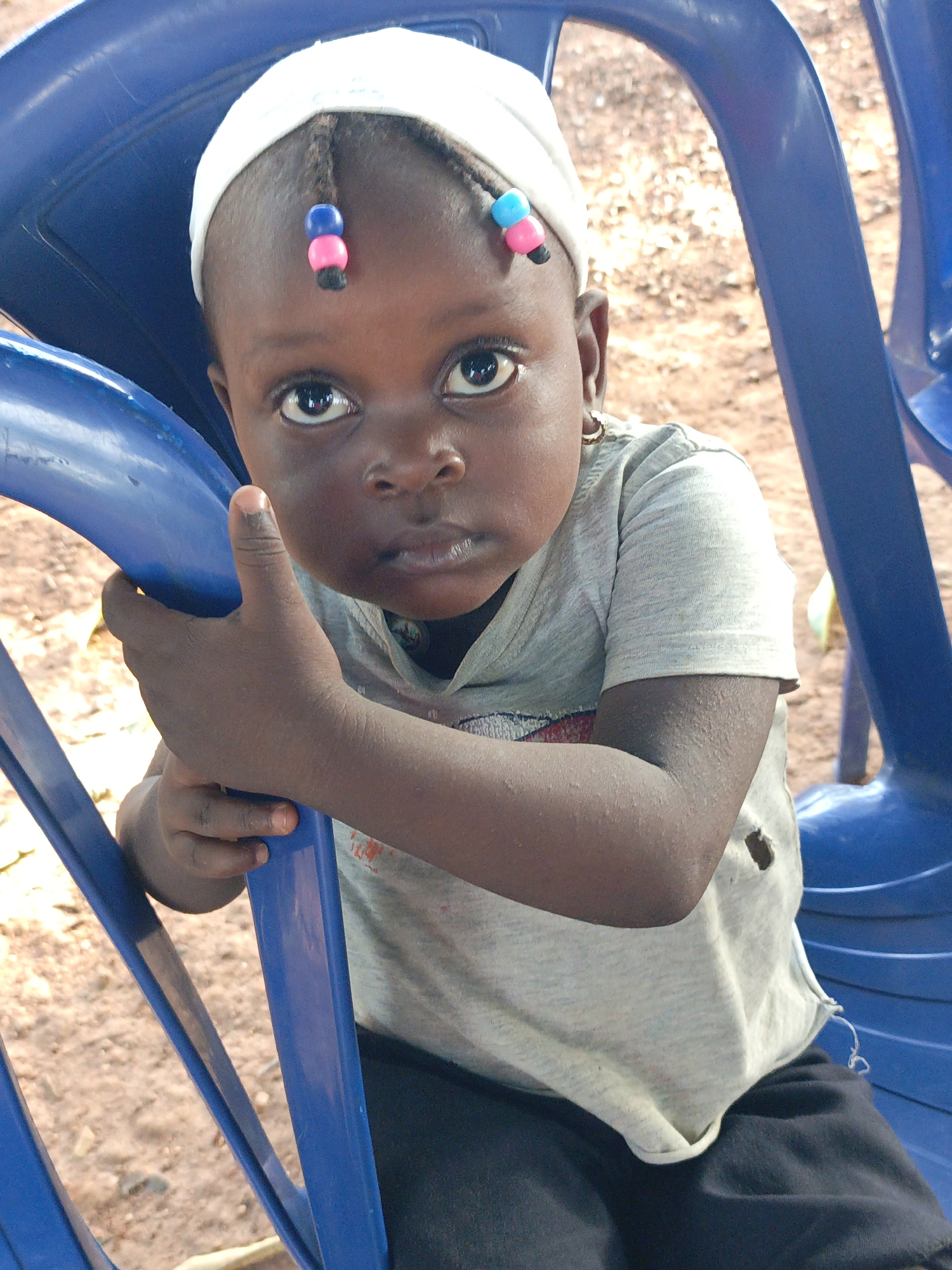 A young child sits in a blue plastic chair outdoors. She wears a white headscarf with two braids adorned with colorful beads. The child has dark skin, large eyes, and a serious expression. Her hands grip the sides of the chair. She wears a light-colored t-shirt and dark pants. The background consists of reddish-brown dirt and a partially visible second blue chair. This image was taken in Akwanga, Nigeria.