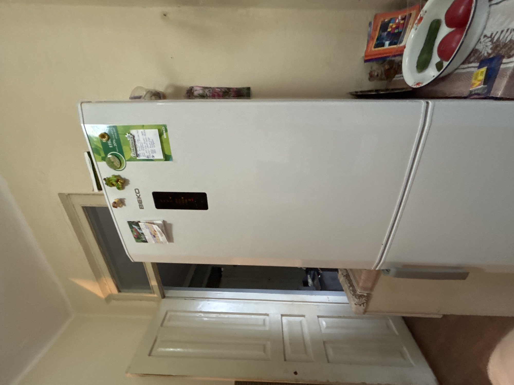 A vertical, medium shot captures a white Beko refrigerator situated in a dimly lit, seemingly residential kitchen. The refrigerator, the primary subject, is adorned with several magnets, including a green circular one featuring text and an image, and two small, cartoonish frog figures. To the left of the refrigerator, a light-colored, double-paneled door is ajar, revealing a dark interior. A portion of another appliance, possibly a freezer or cabinet, is visible to the left of the refrigerator. The wall behind the refrigerator is painted a pale yellow. On the right side of the refrigerator, a table is set with a white oval bowl containing a green cucumber and two red tomatoes. Behind the bowl, stacked books or photo albums are visible, along with a small glass jar. The flooring is a dark wood. The overall impression is one of a lived-in, somewhat cluttered domestic space.