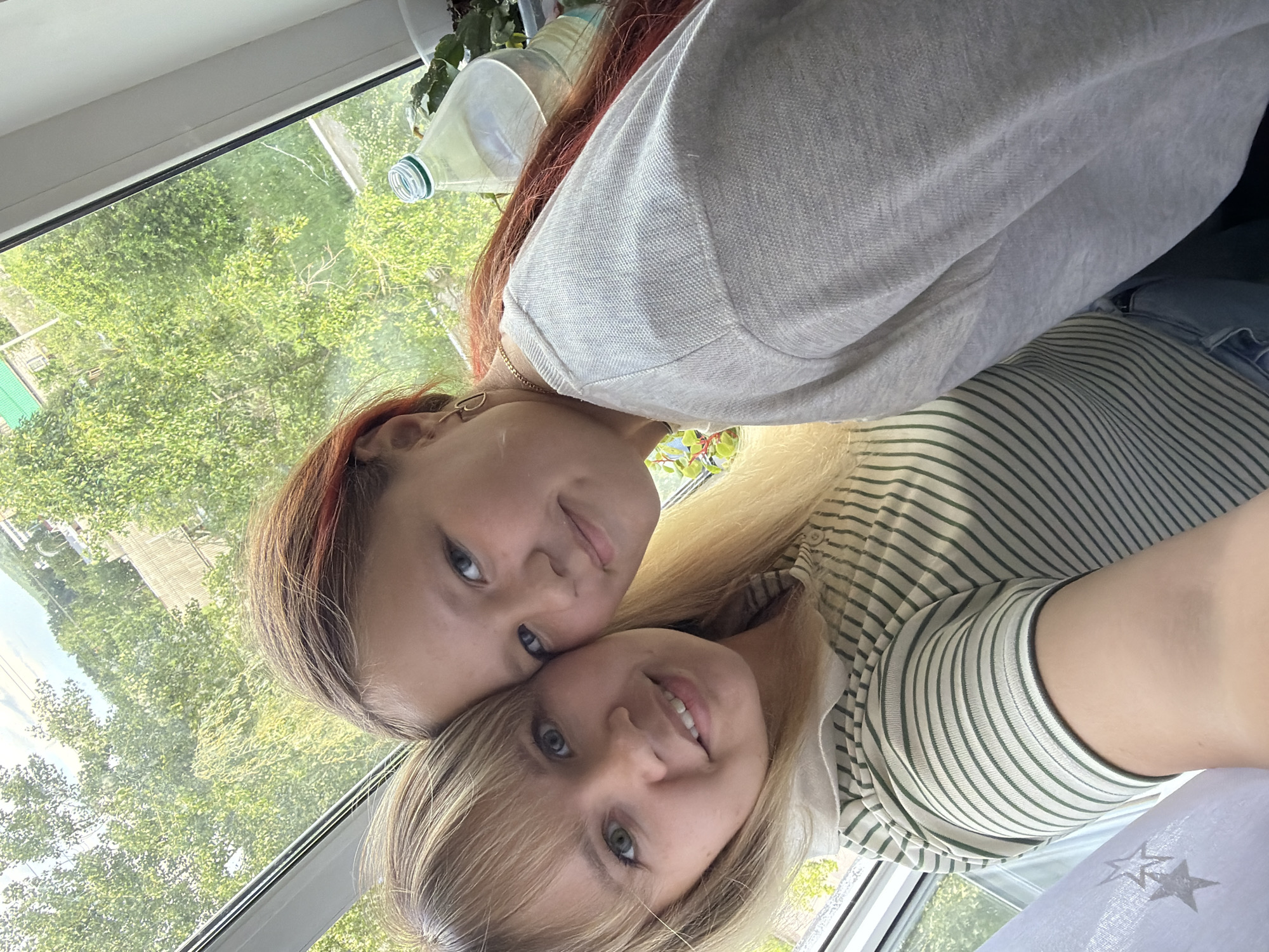 Two females pose close together. The first female has auburn and brown hair, fair skin, and blue eyes. She wears a light grey long-sleeved shirt and a gold chain necklace. The second female has blonde hair, fair skin, and blue eyes. She wears a white shirt with green stripes. A white lace fabric is in the lower right corner. A bottle and green foliage are behind them. In the background is a view through a window with green foliage and buildings. Based on metadata, the image was likely taken in the city of Chaikovsky, Russia.
