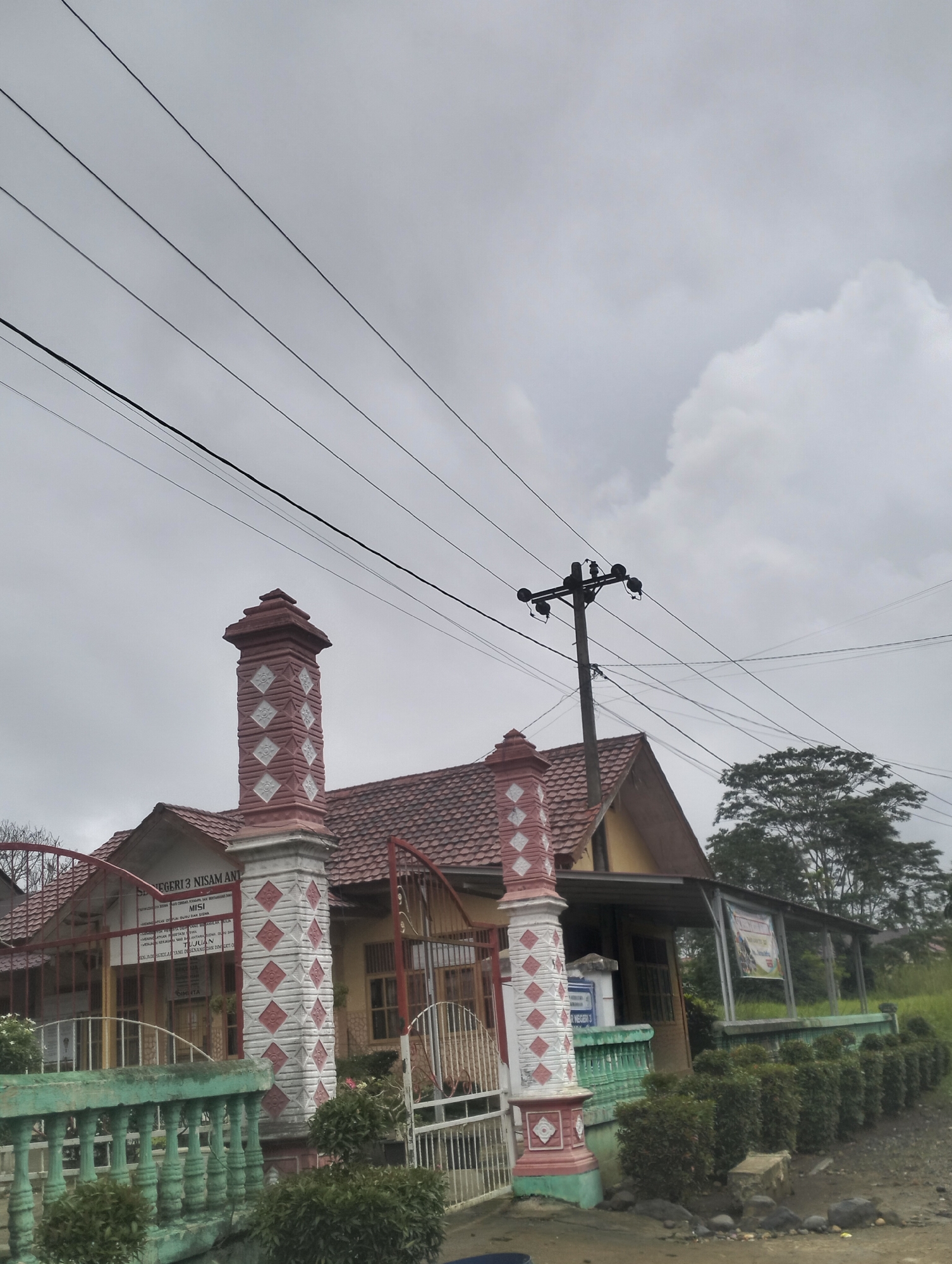 The image shows the entrance to a building, possibly a school, with a red-tiled roof and yellow walls. A decorative gate with red and white diamond patterns is visible, along with a green-painted fence. Overhead, multiple power lines stretch across a cloudy sky, supported by a utility pole. In the background, trees and vegetation are present. A sign above the entrance reads "SEKOLAH NEGERI 3 NISAM ANTARA". The overall scene depicts a building exterior in a possibly rural or suburban setting, with infrastructure elements such as power lines indicating an inhabited area. The image appears to be taken from a slightly elevated, ground-level perspective looking upwards towards the building and the sky.
