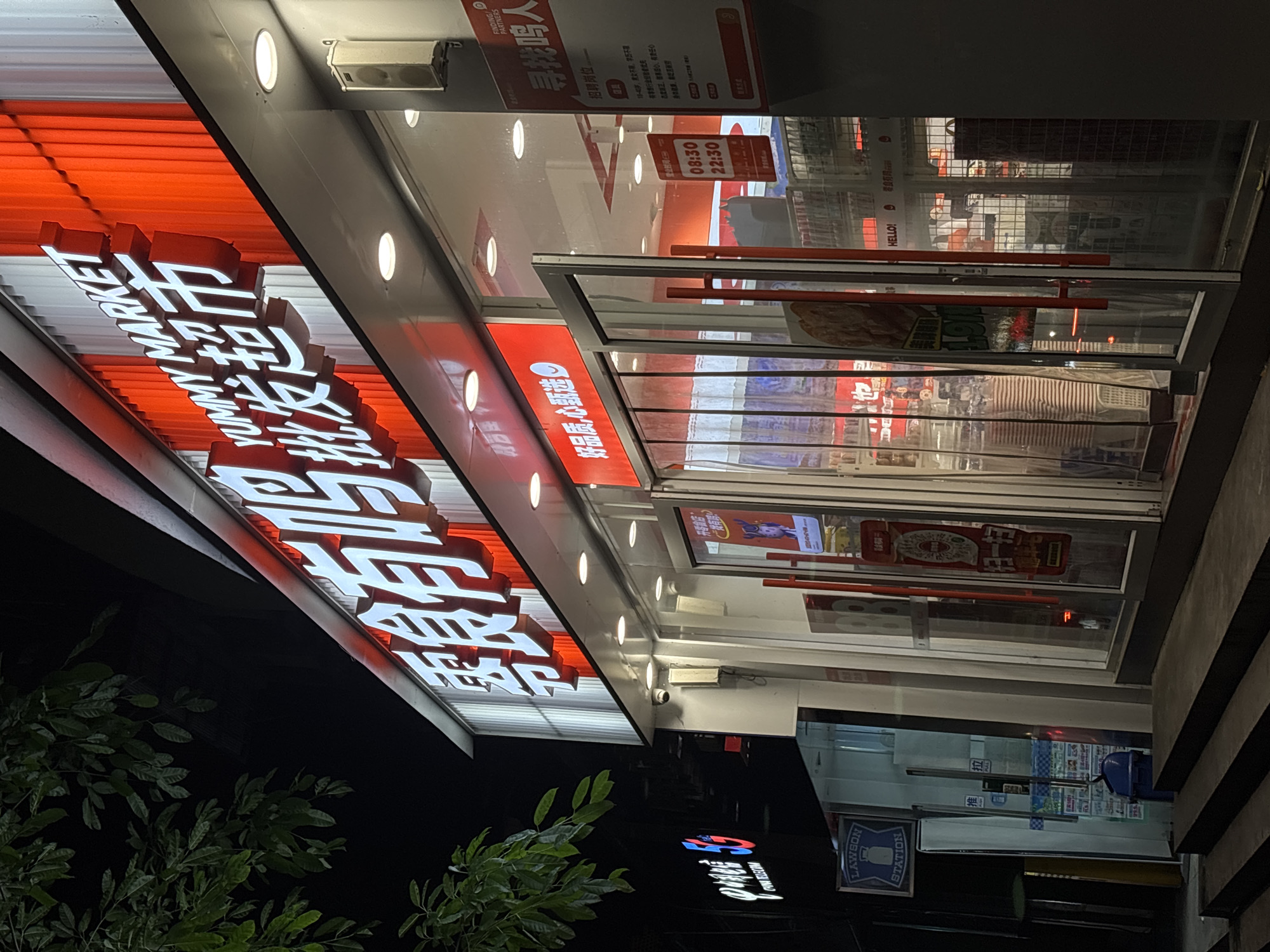 The image captures a storefront in Nan'an District, China. The illuminated sign reads "Yummy Market," adjacent to a Lawson Station store. The storefront features opening hours of 08:30 to 22:30, and recruitment notices for staff. The environment includes green foliage visible on the left.