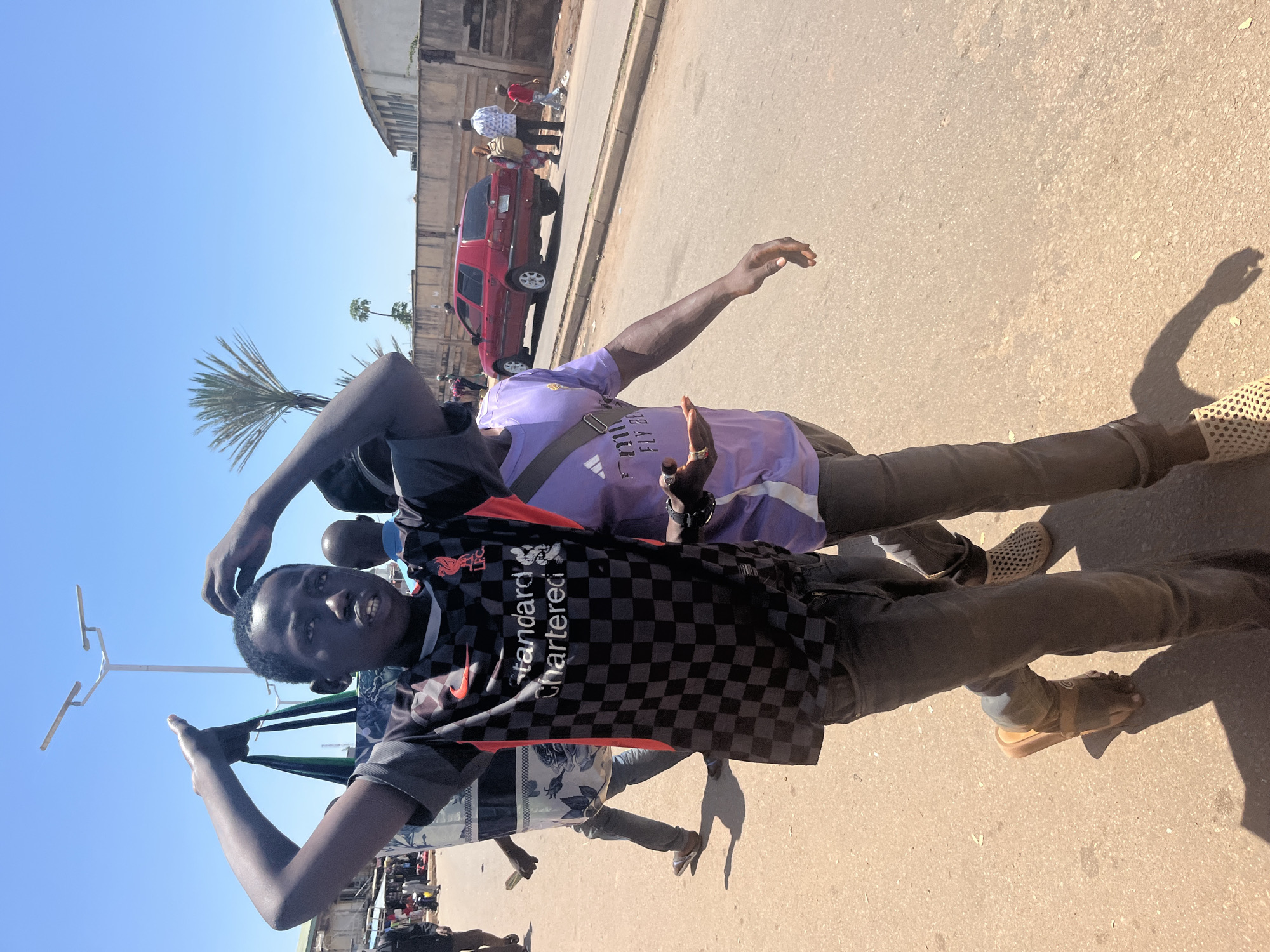 Two young men walk along a dusty street under a bright blue sky in Jos, Nigeria. The man in the foreground on the left wears a black and grey checkered Liverpool FC jersey with "Standard Chartered" printed across it, and dark grey jeans. He holds his hands up to his head, adjusting his hair or shielding his eyes from the sun. He has a dark complexion and is wearing brown sandals. To his right, a second young man walks with his left arm extended, wearing a light purple t-shirt with "FLY BY" visible on it, and dark grey jeans. He also appears to have a dark complexion. In the background, a red SUV is parked on the side of the road, and several other people, including adults and a child, are visible walking in the distance. Buildings line the right side of the street, and a tall palm tree is visible against the sky. The ground is a light brown, dusty surface, suggesting a dry environment. A street light pole with multiple solar-powered lamps is visible in the upper left portion of the image.