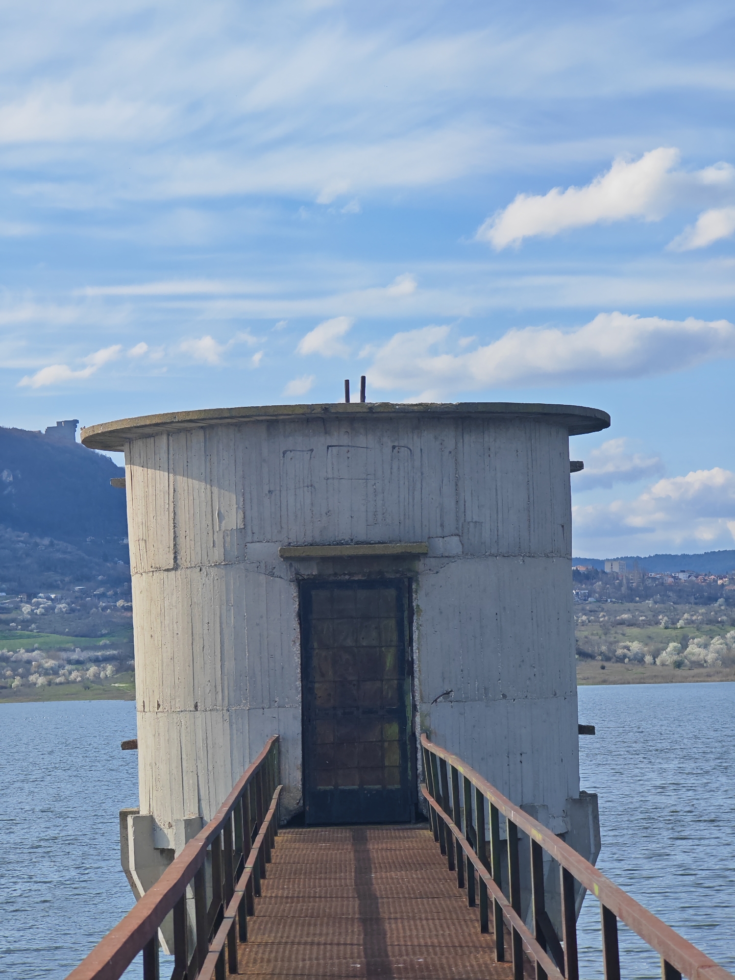 The image depicts a cylindrical concrete structure with a flat roof, situated in the middle of a body of water, likely a lake or reservoir. A metal walkway with rusty railings leads up to a metal door in the structure. The sky is blue with scattered white clouds. In the background, there is a landscape with trees, possibly a small village or town, and a mountain ridge. This specific structure is located in the city of Shumen, Bulgaria.