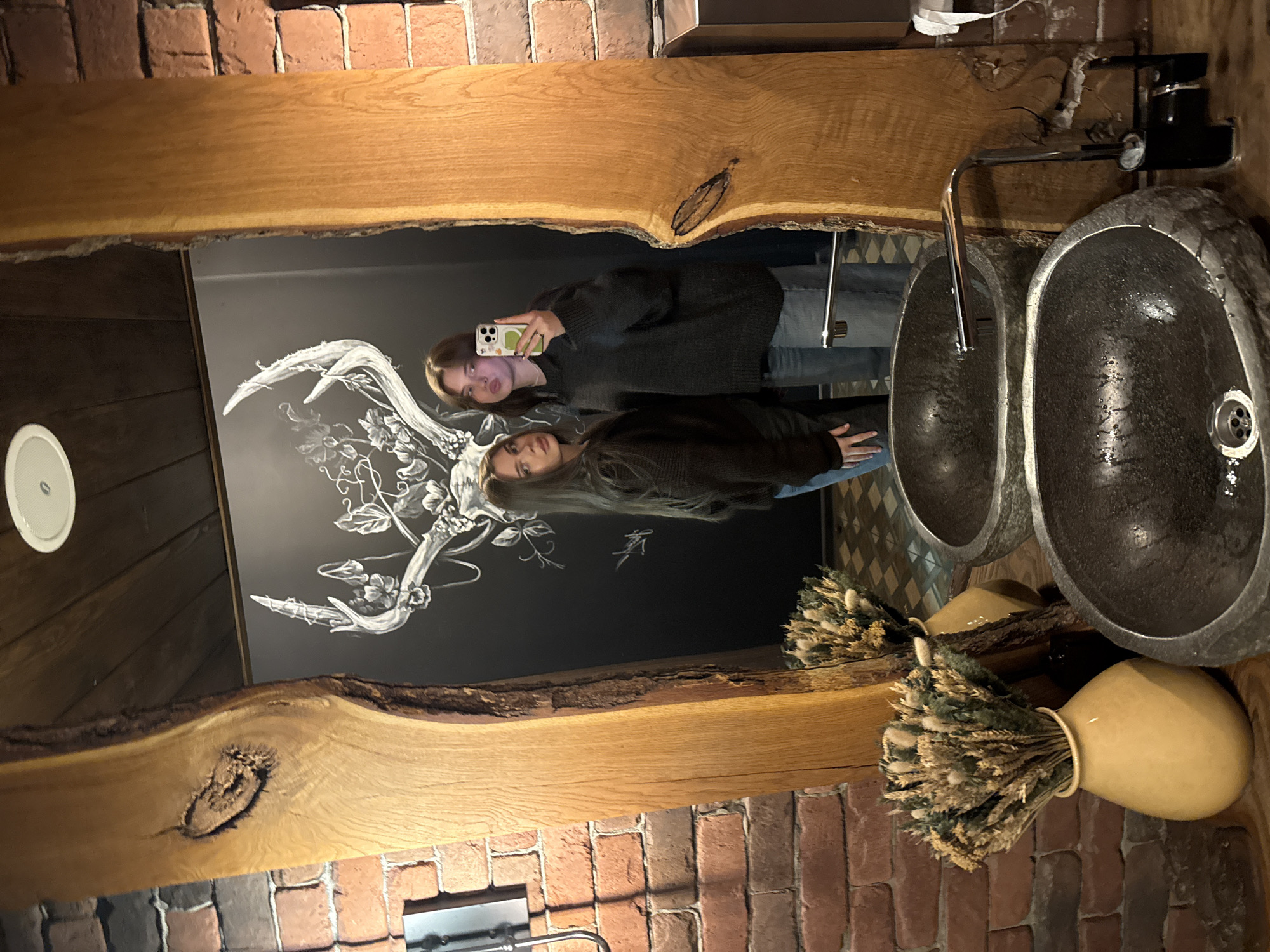 A selfie is captured in the reflection of a large, rough-edged wooden-framed mirror above a double-sink vanity in a room with brick walls. A black and white drawing of antlers decorates the wall behind the mirror. Two young women are reflected in the mirror, with one holding a smartphone up to take the picture. The vanity has two stone-look sinks, each with a chrome faucet. Beside one of the sinks sits a tan-colored vase filled with dried plant material. The image was taken in the Krasnoyarsk, Russia.