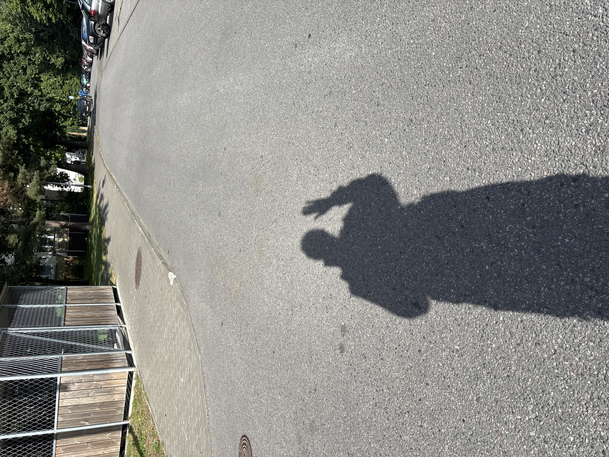 The image depicts a shadow of a person on an asphalt road in Tallinn, Estonia. The person's shadow is cast by the sun and shows the silhouette of a person with their right hand raised in a waving gesture. The shadow is tall and elongated, indicating the sun is relatively low in the sky. The background consists of a paved sidewalk with a metal fence and wooden panels bordering a grassy area on the left. Further down the road, parked cars and trees are visible, suggesting a residential or urban environment. The road surface is dark grey asphalt with visible aggregate. The lighting suggests it is daytime, likely late afternoon or early morning.